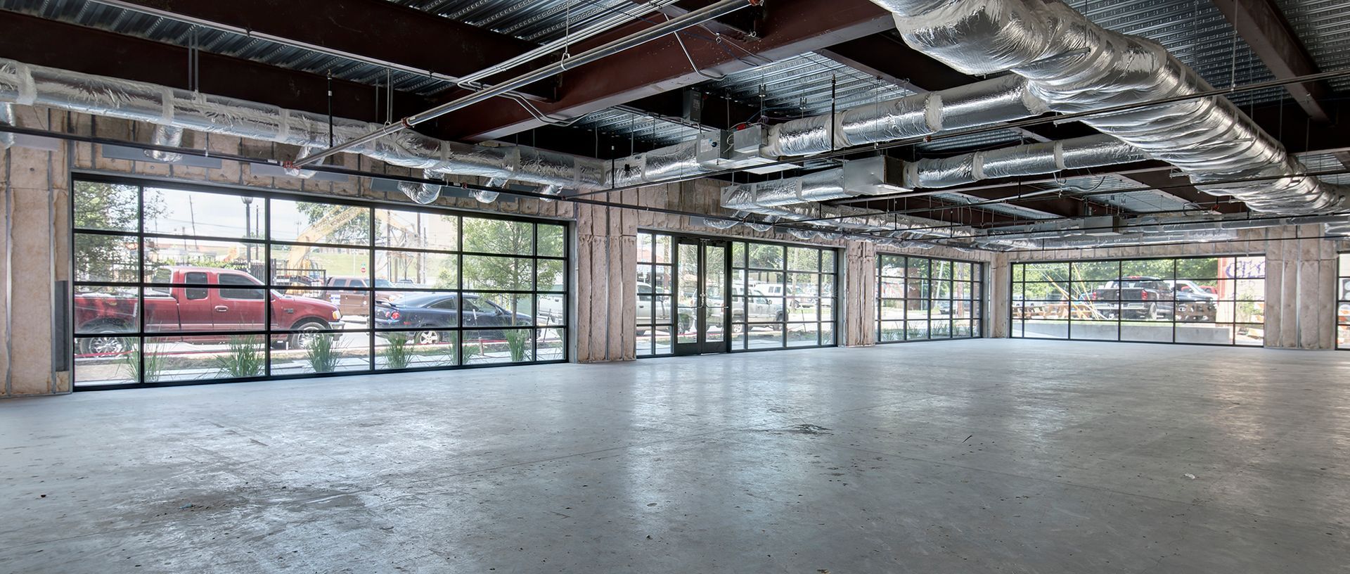 An empty open-plan building with large glass windows showing the exterior and ceiling ducts.