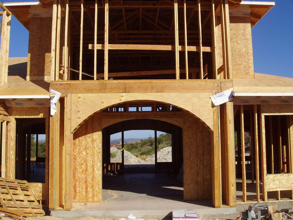 Wooden house under construction, arched entryway, framing visible, blue sky, distant view.