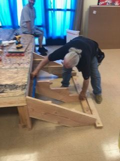 Two men building a wooden structure indoors, one uses a level, another observes.