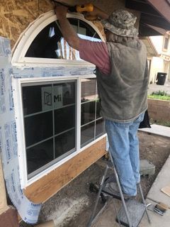 Man on ladder installing a window. He is wearing a hat, vest, and jeans. The house is under construction.