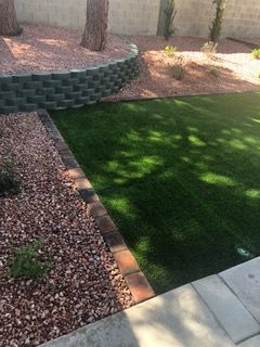 Lush green grass area bordered by a red brick border, next to brown gravel and retaining wall.