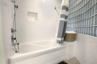 White bathroom with tub, shower, gray and white striped curtain, glass block wall.
