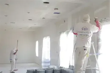 Painters in white overalls spraying walls and ceiling in a bright, empty room.