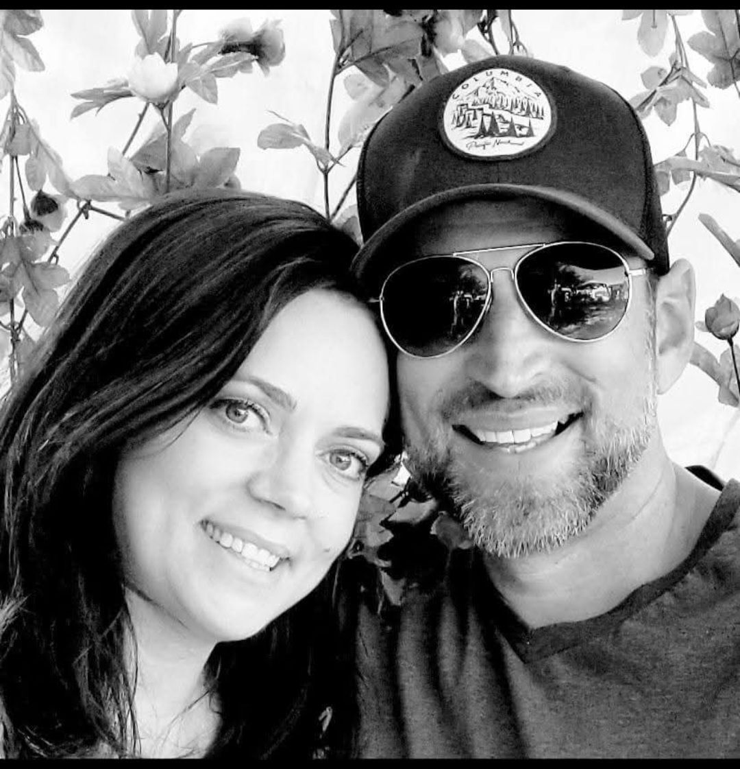 Smiling couple posing close together; woman with dark hair, man in sunglasses, wearing a hat.