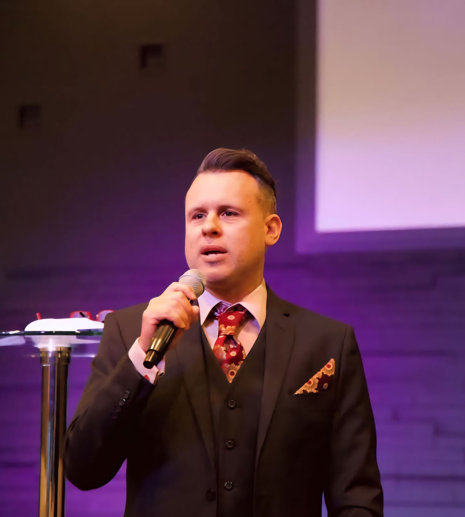 Man in suit holding a microphone, speaking at a podium in a room with a purple background.