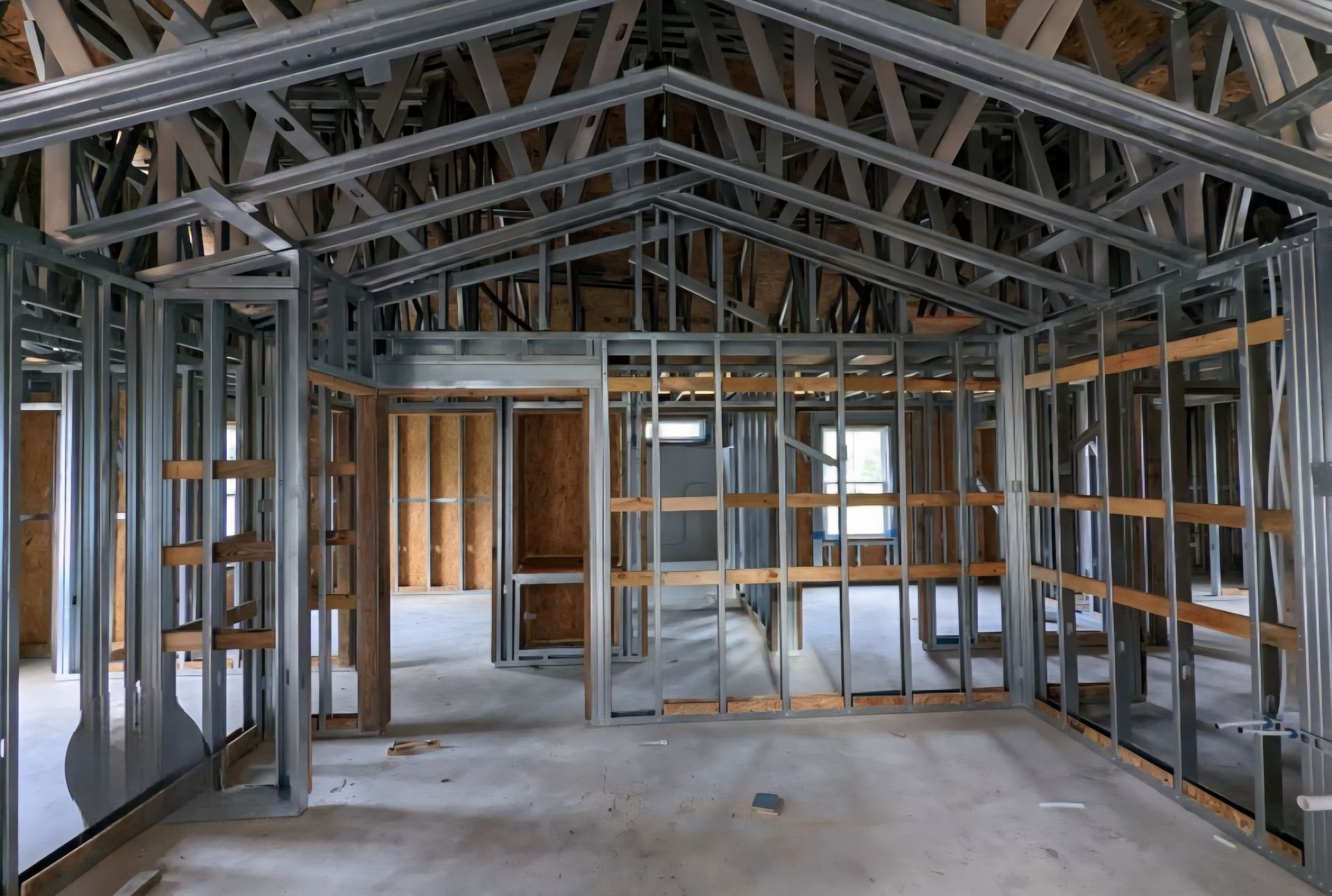 Interior construction view: Metal framing of a house's walls and roof, with some wooden studs, cement floor.