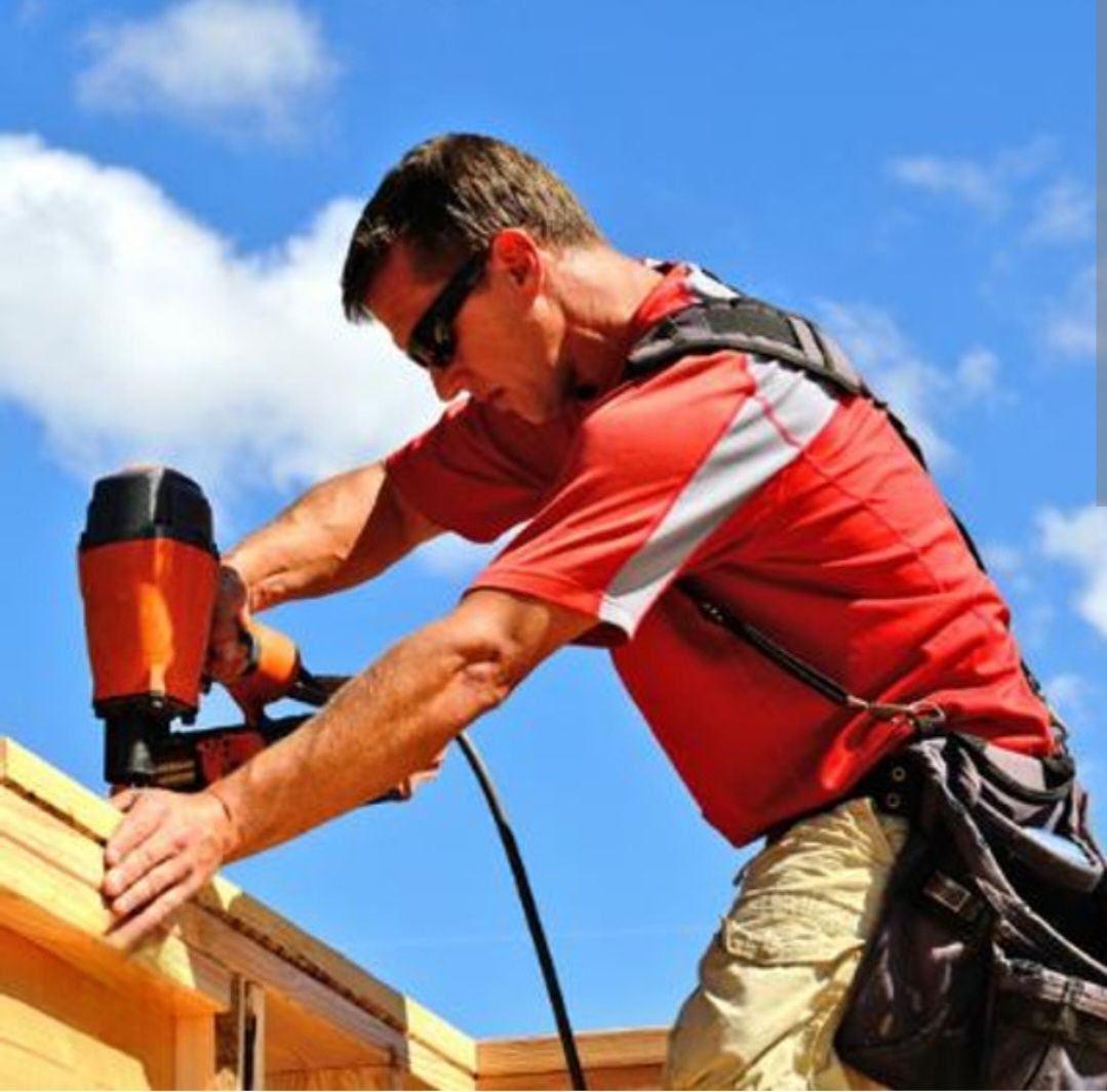 Construction worker using a nail gun on wooden frame, wearing sunglasses and red shirt, outdoors.