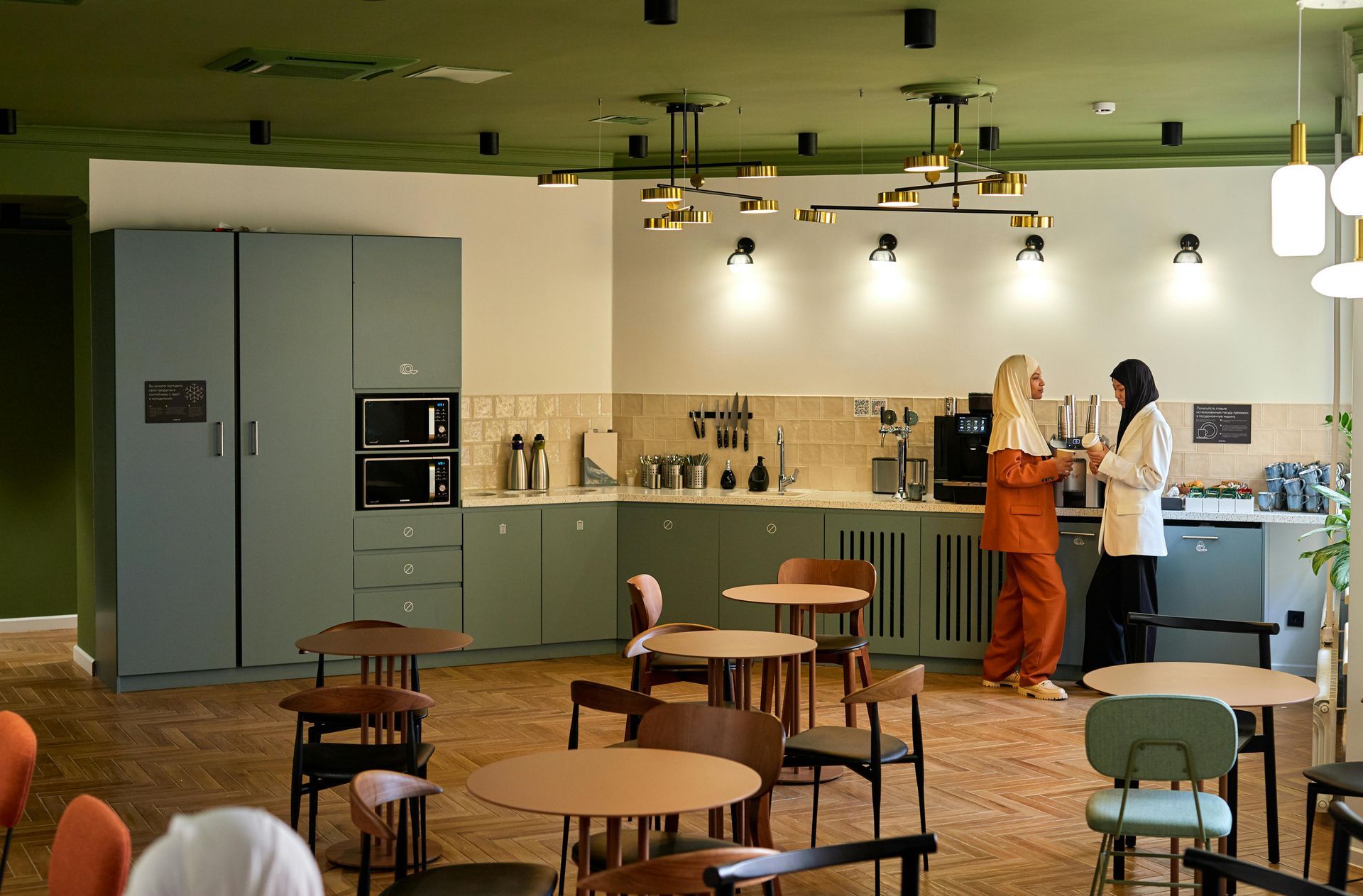 Two women in a green-themed breakroom, near the coffee station, chatting. Tables and chairs fill the room.