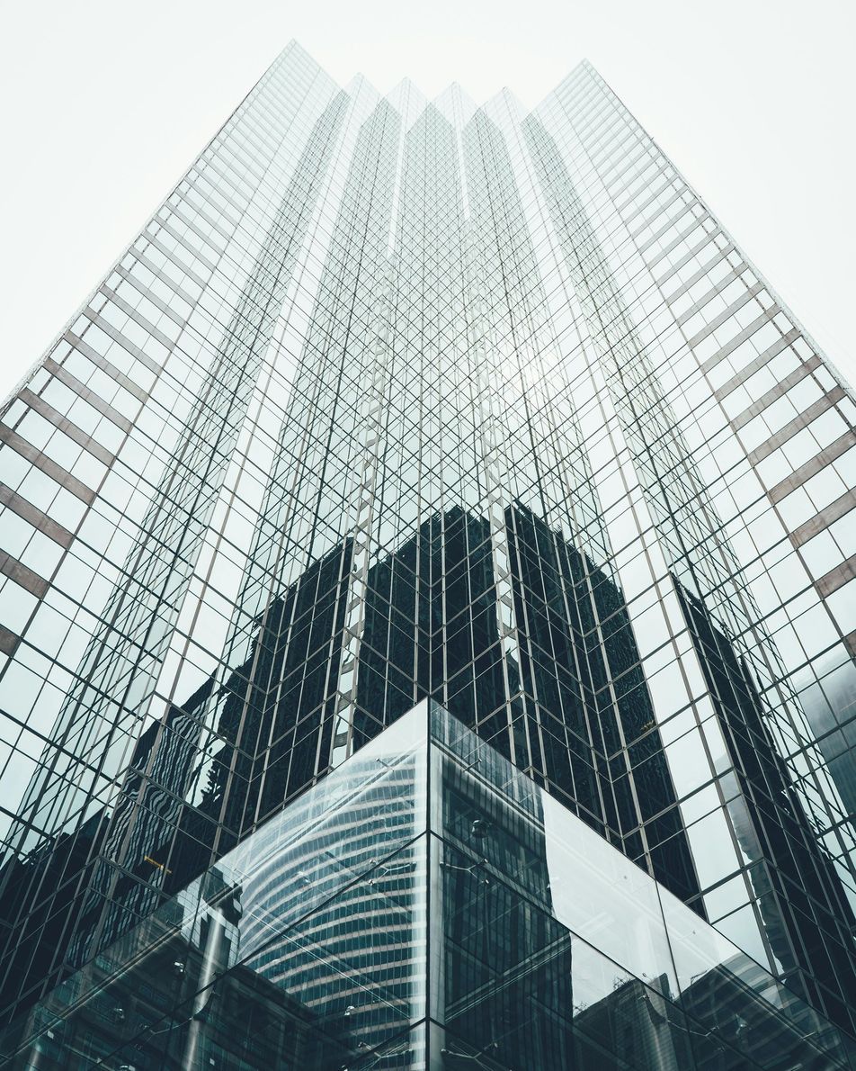 Tall glass skyscraper, viewed from below. Reflections of the sky and surrounding buildings in the windows.