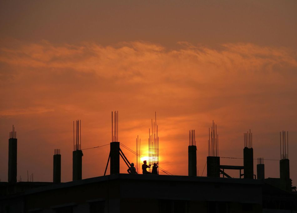 Silhouetted construction workers on a building's rooftop at sunset, against an orange sky.