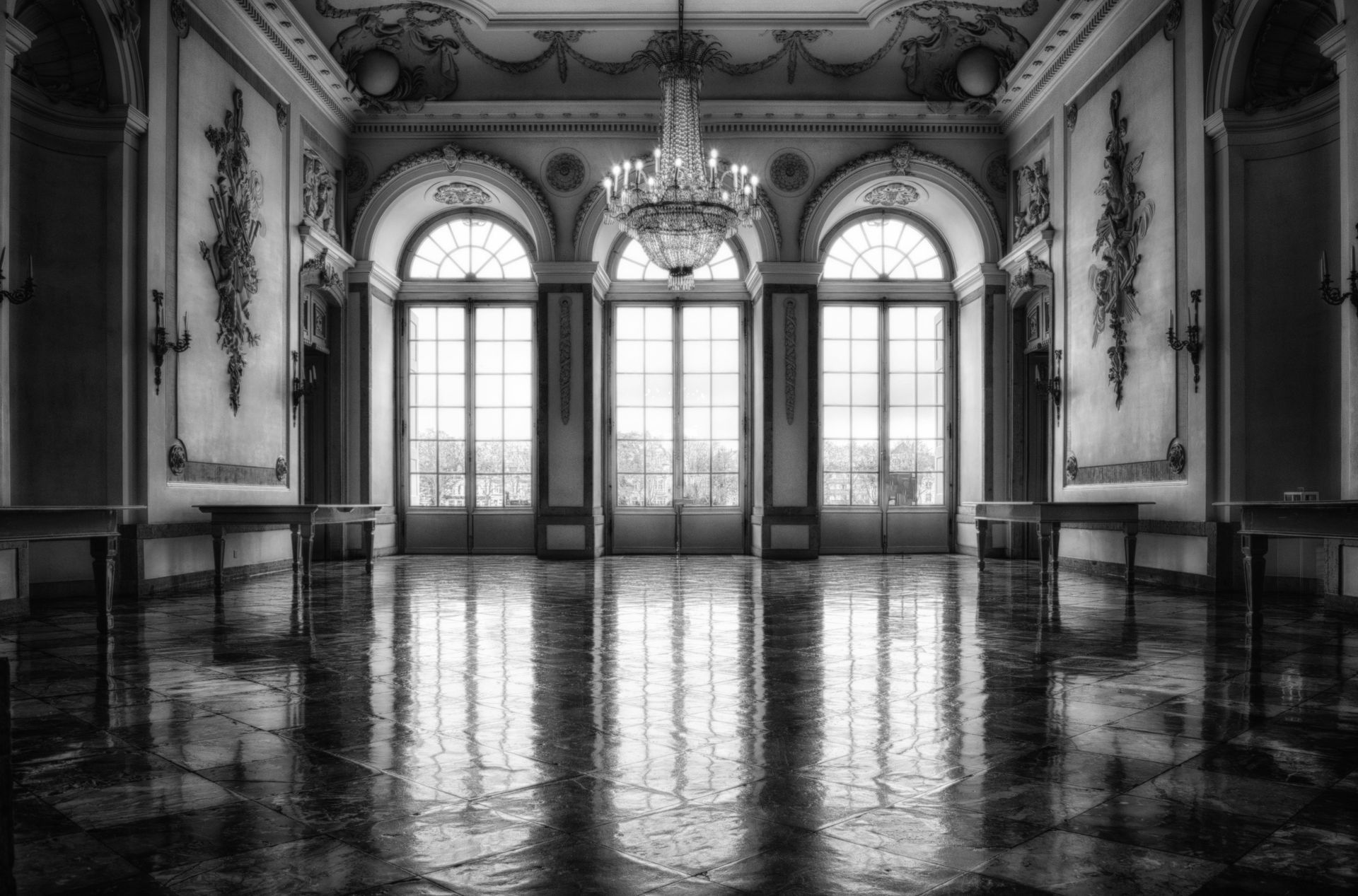 Elegant hall, black and white. Crystal chandelier hangs above mirrored floor and tall windows.