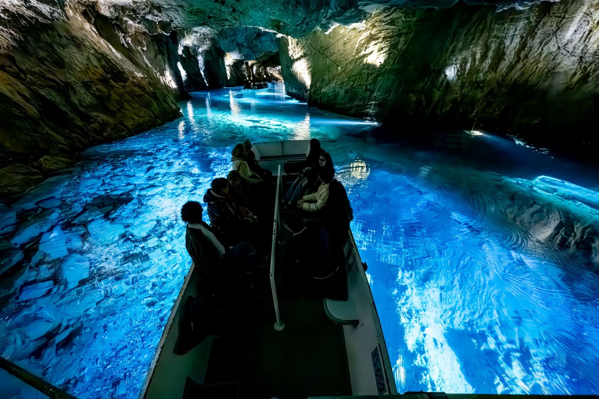 Boat of people in a glowing blue cave. Dark rocks surround the water.