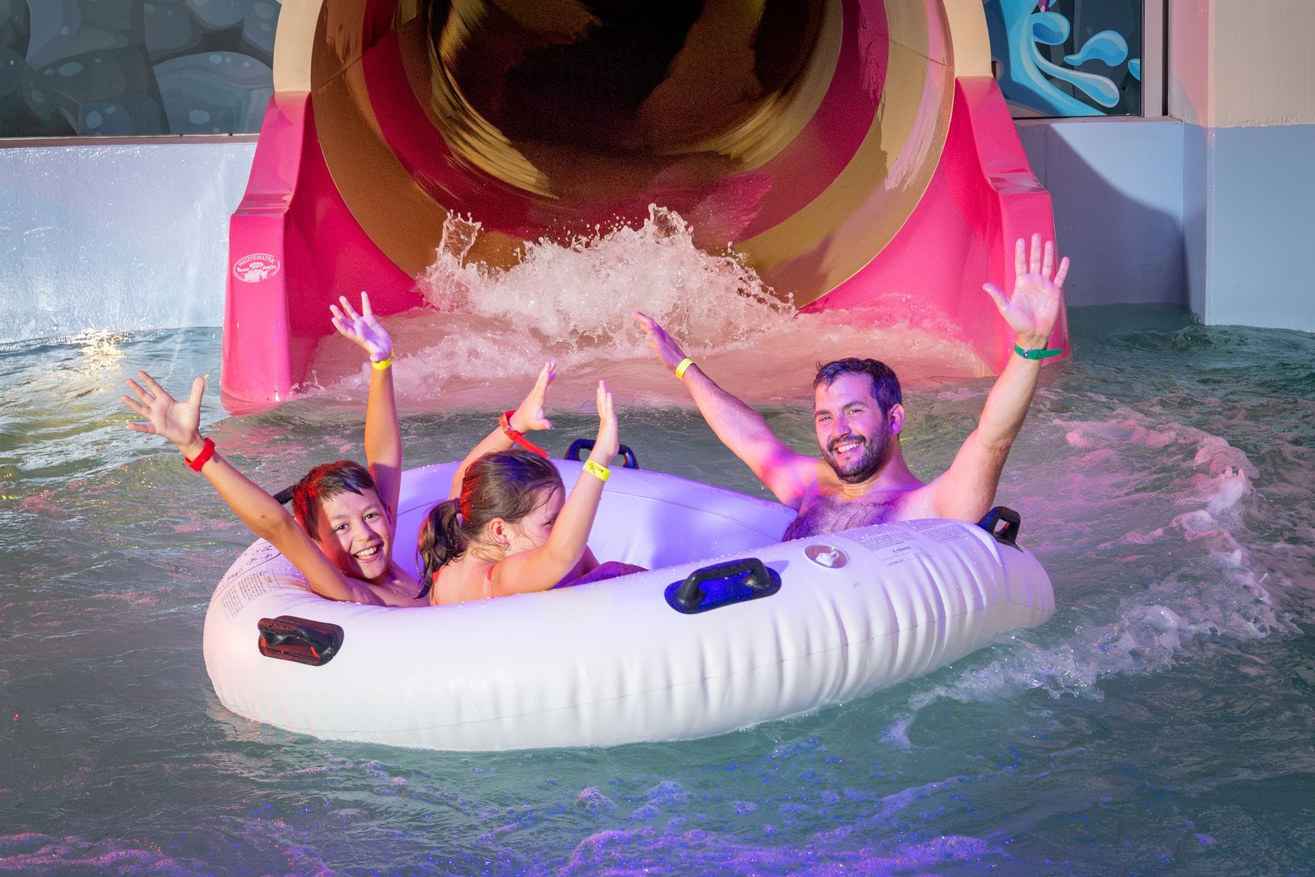 Family on inflatable raft exiting water slide, raising arms in excitement. Pink and tan slide, indoor pool.