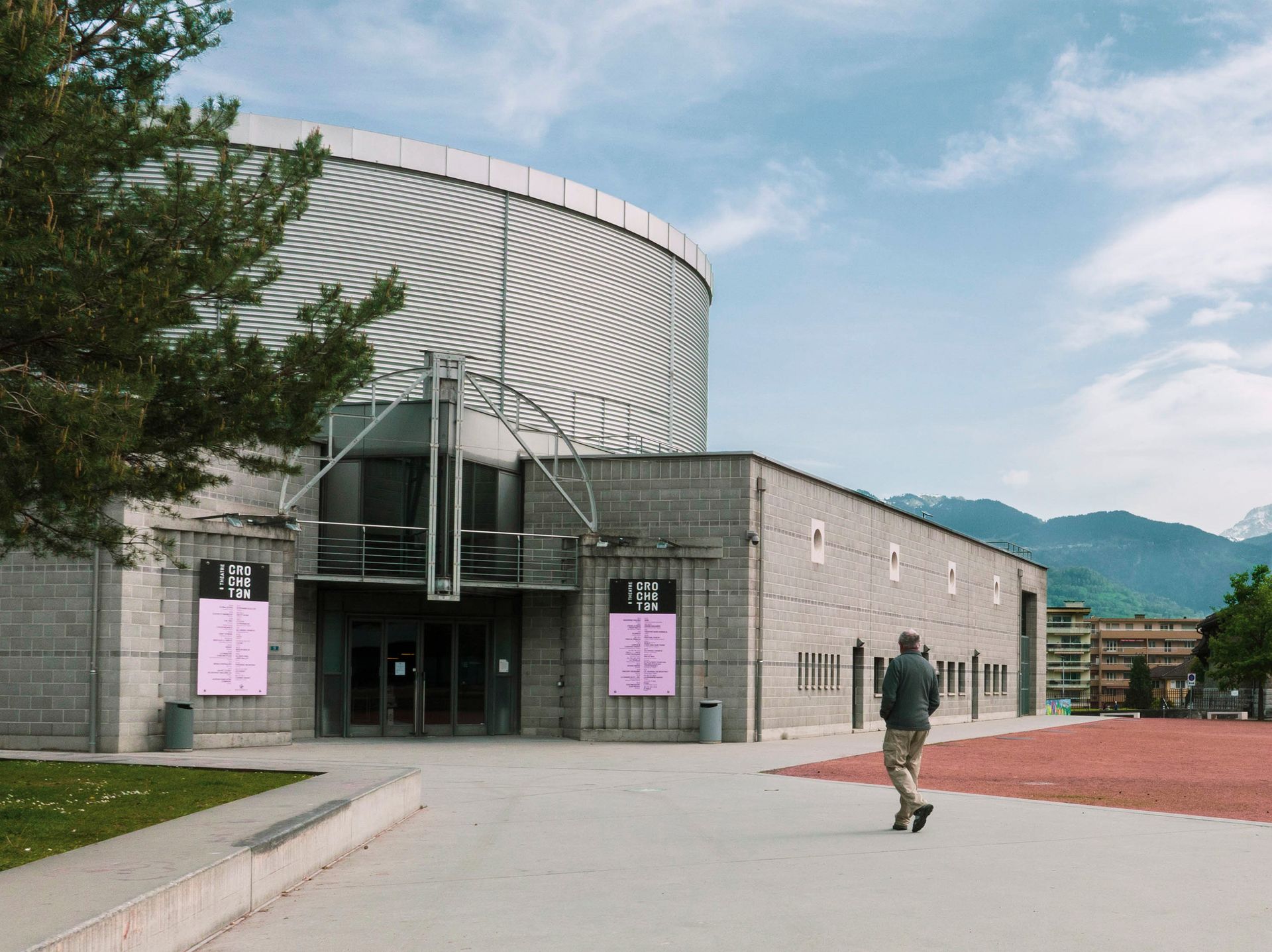 A man walks toward a modern building with a patterned metallic top and posters on the facade; blue sky.