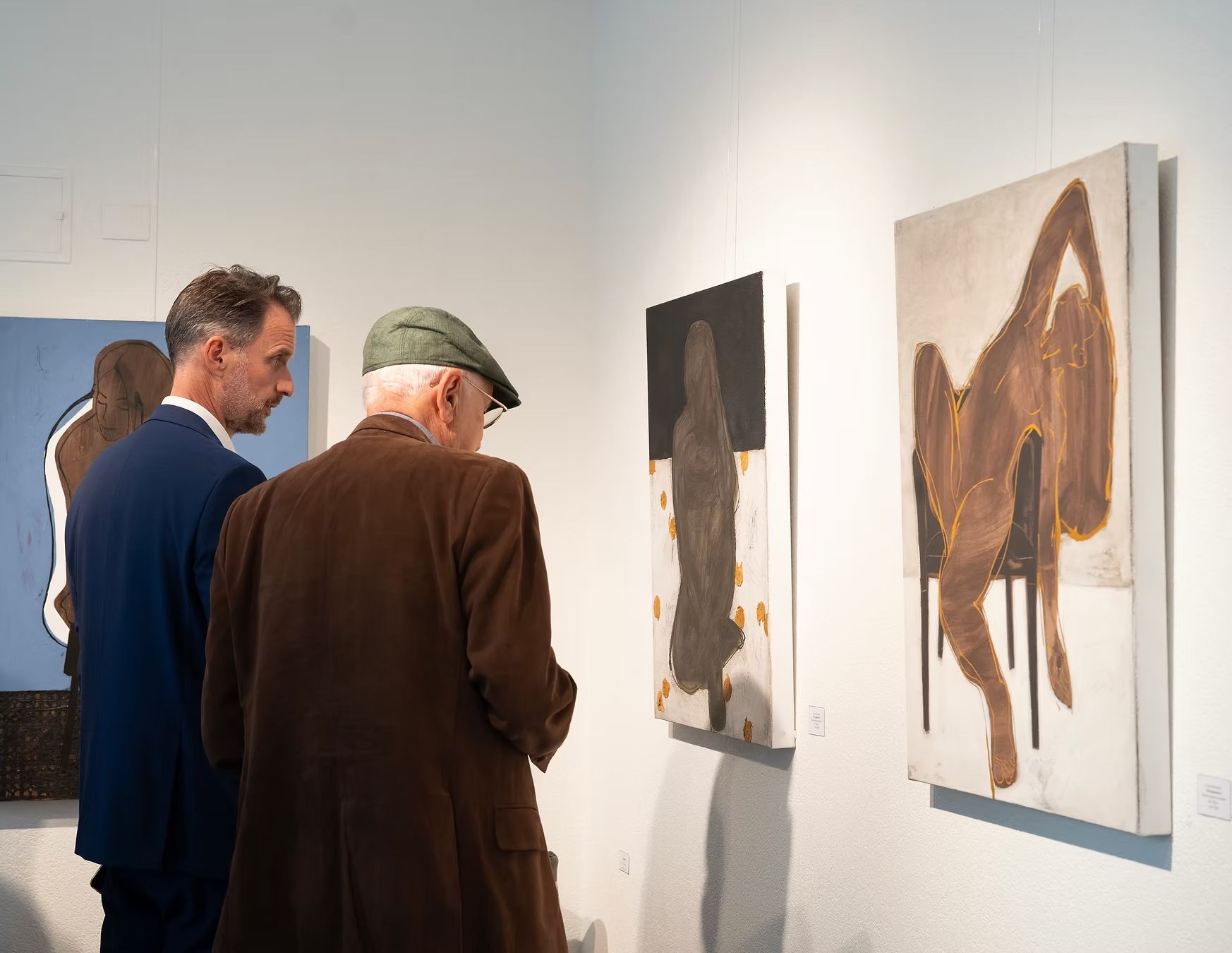 Two men at an art gallery, viewing abstract paintings on a white wall.