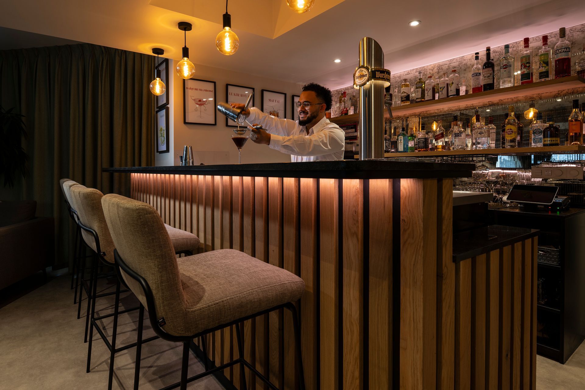 Bartender pouring drink at a lit-up bar. Three bar stools in front. Wooden paneling, bottles on shelves.