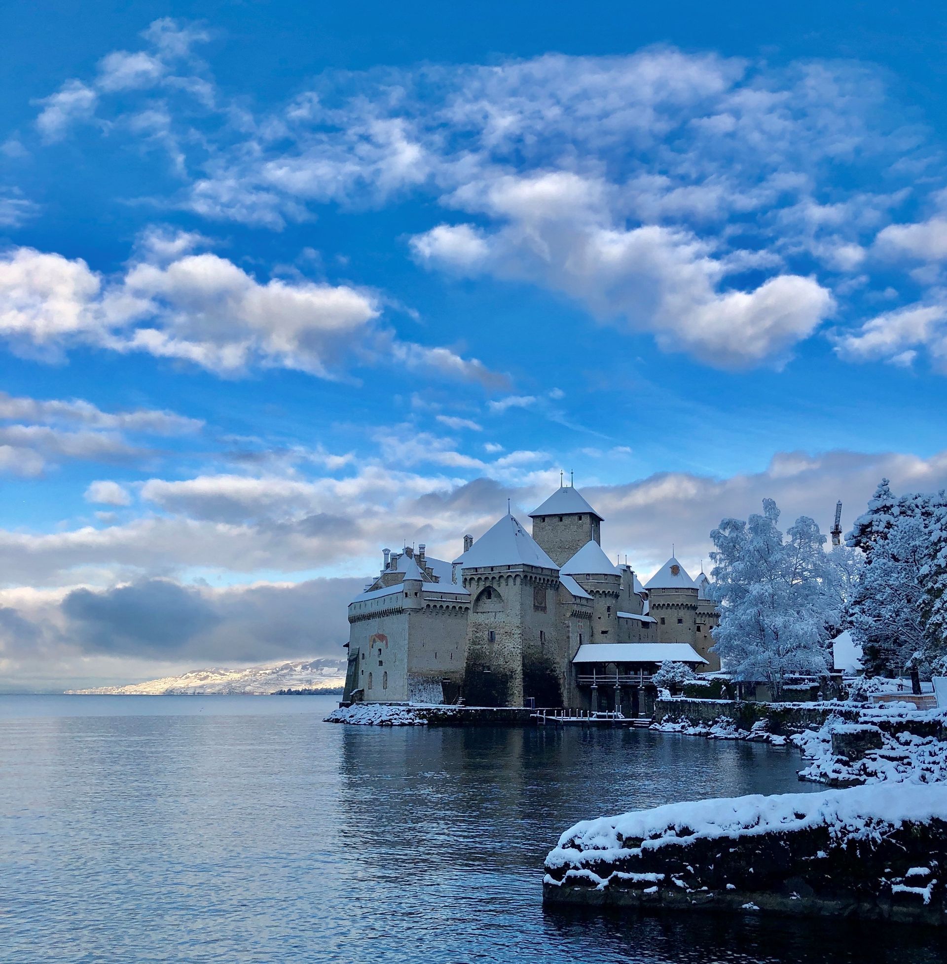 Snow-covered Chillon Castle on Lake Geneva under a partly cloudy blue sky.