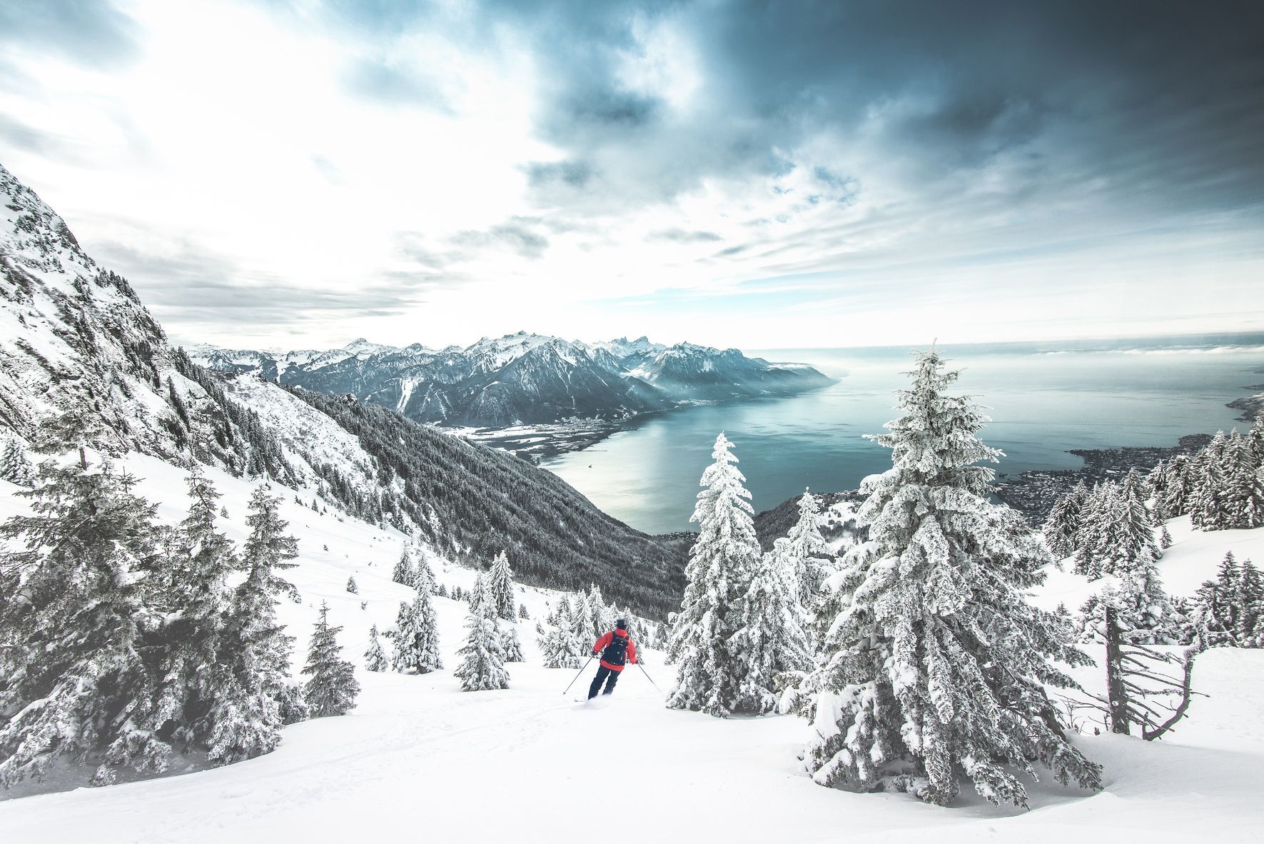 Skier descends snowy mountain slope; snow-covered trees, lake, and mountains in the background under a cloudy sky.