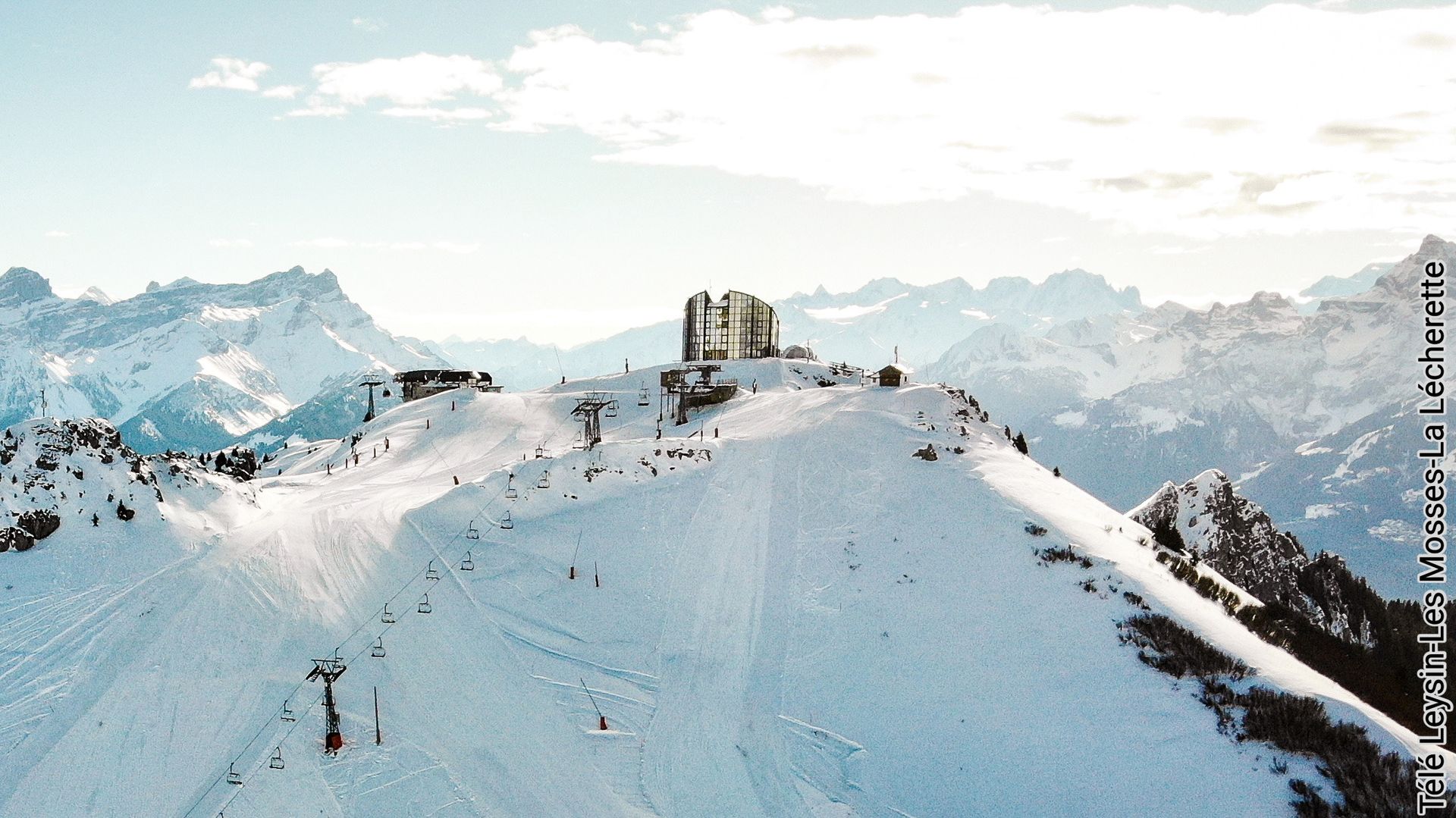 Snow-covered mountain peak with a building and skiers, against a backdrop of snowy peaks and a blue sky.