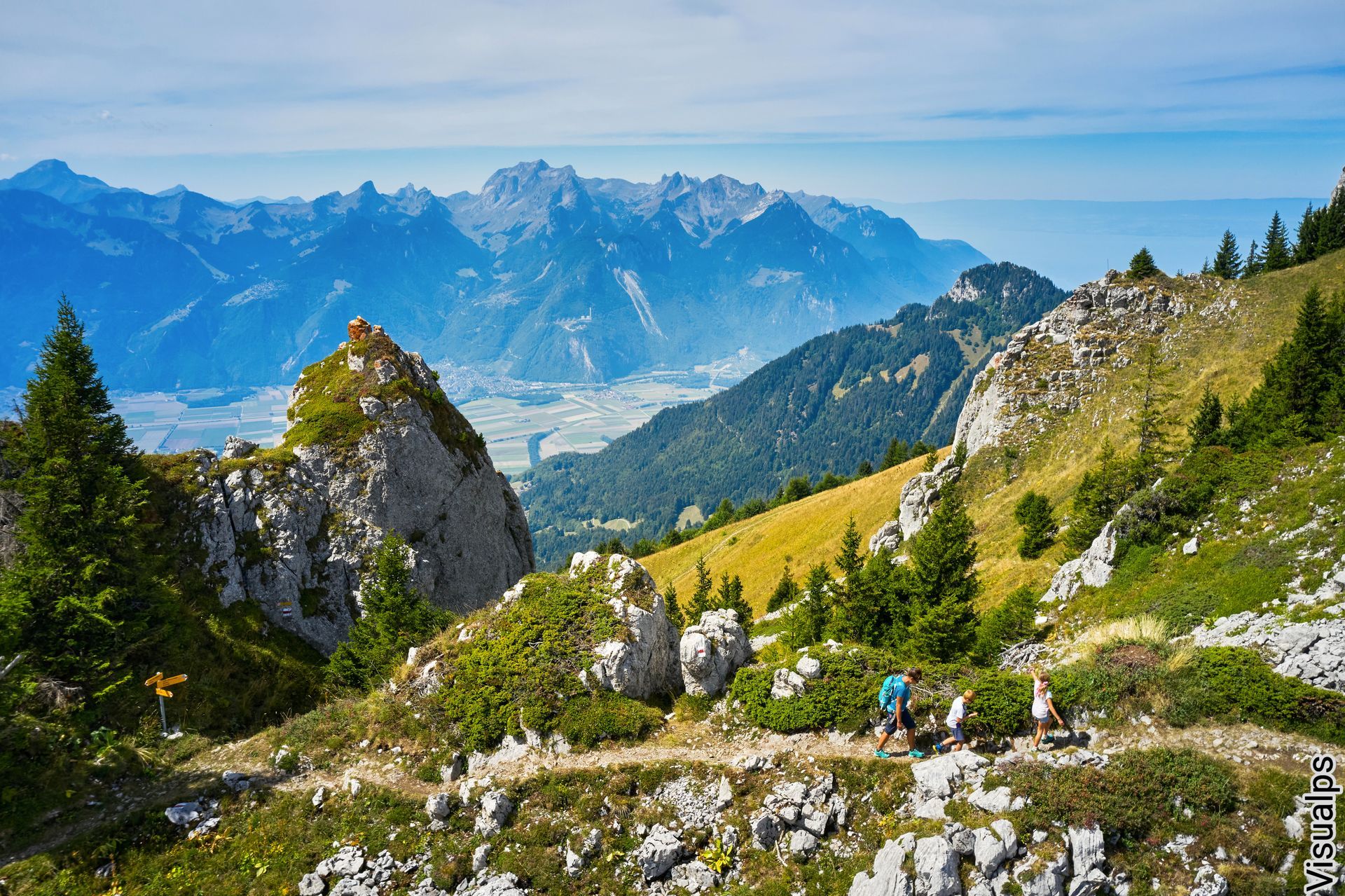 Mountain trail with rocky peaks, green meadows, and distant blue mountains under a clear sky