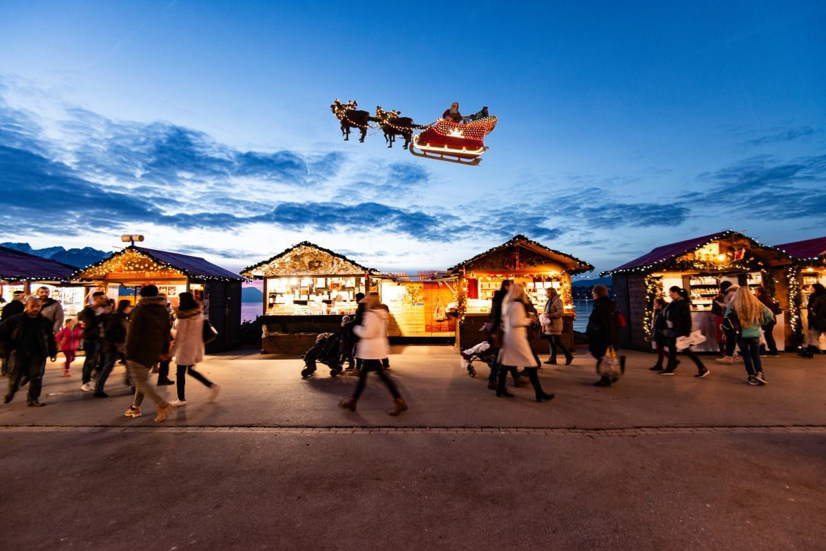 Santa's sleigh flies over a Christmas market with wooden stalls and people; twilight sky.