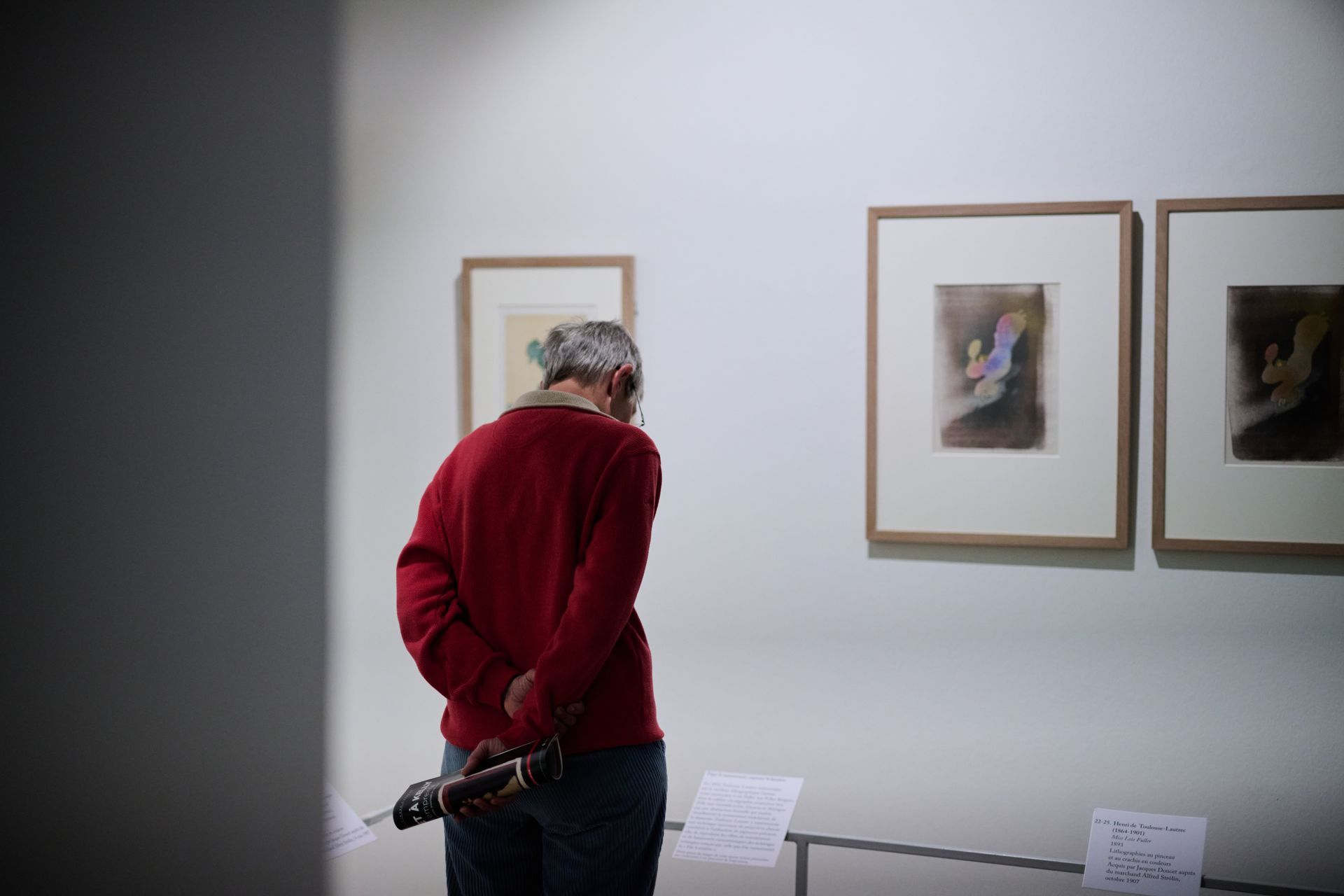 Person in red sweater looks at framed artwork in a gallery.
