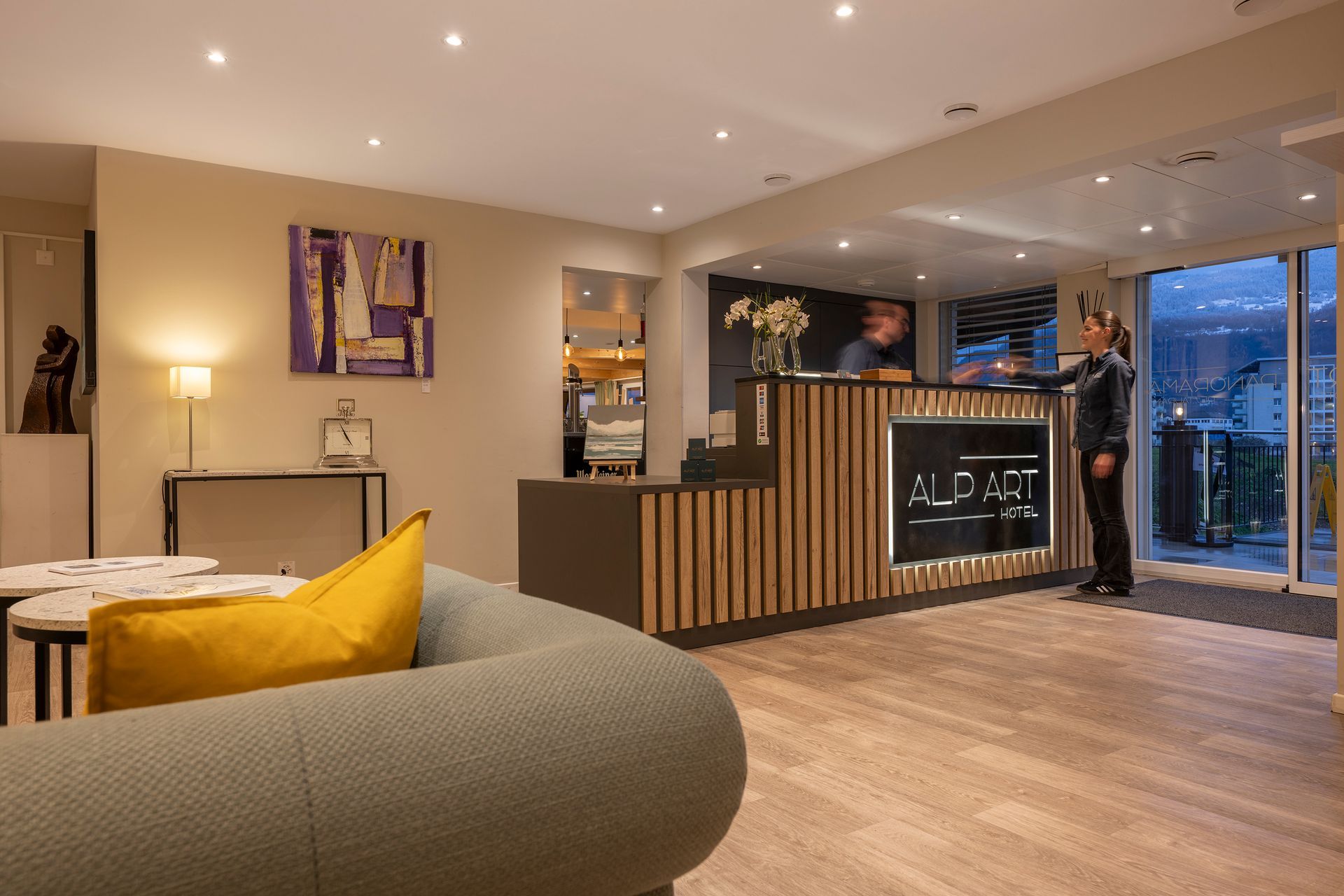 Réception d'hôtel avec un employé au comptoir. Éléments en bois, mobilier moderne et une personne debout près de l'entrée.