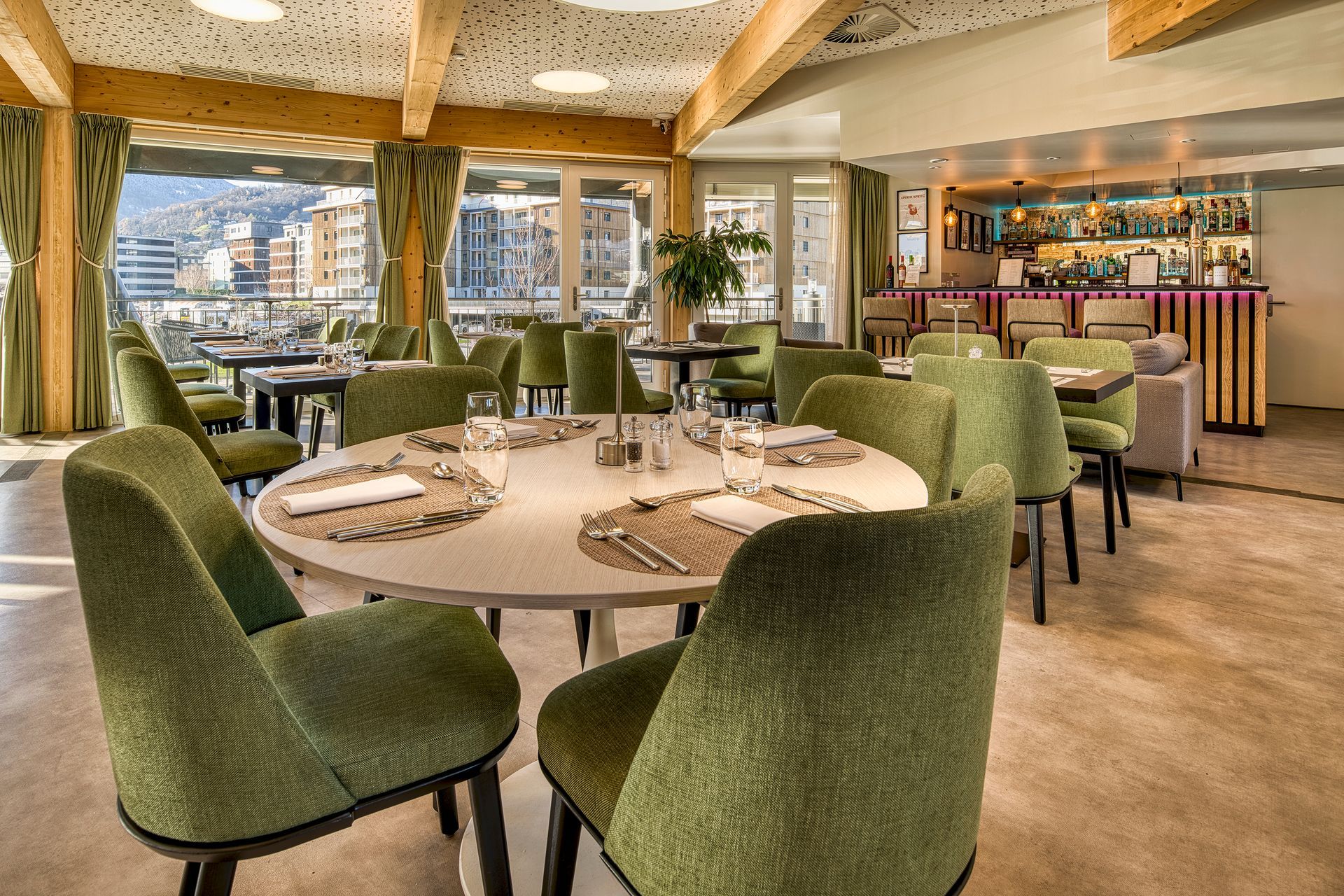 Interior of a restaurant with green chairs, round tables set for dining, and a bar in the background.
