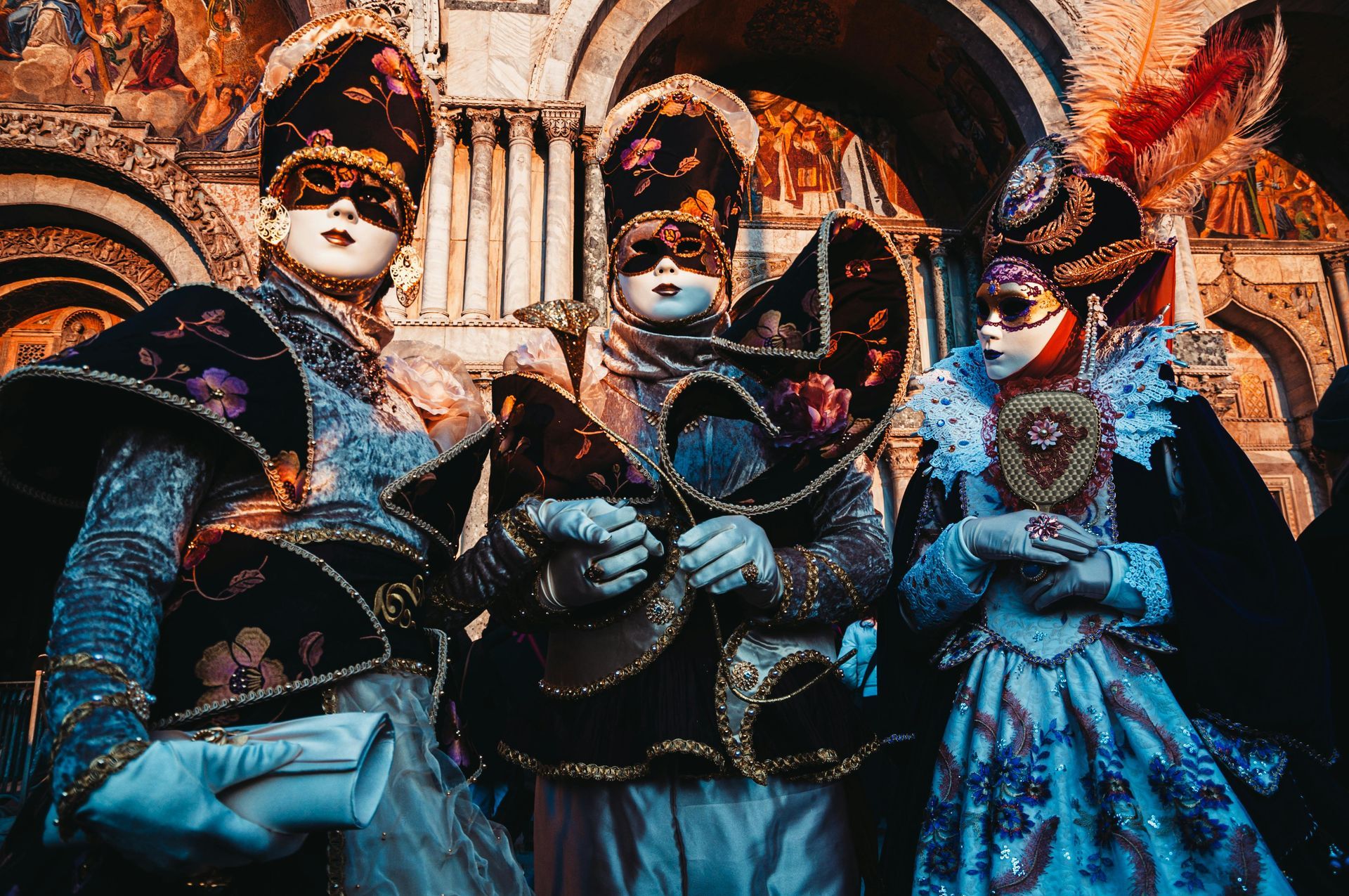 Three people in ornate Venetian Carnival costumes, masks, elaborate hats, and gowns, posing in front of an archway.