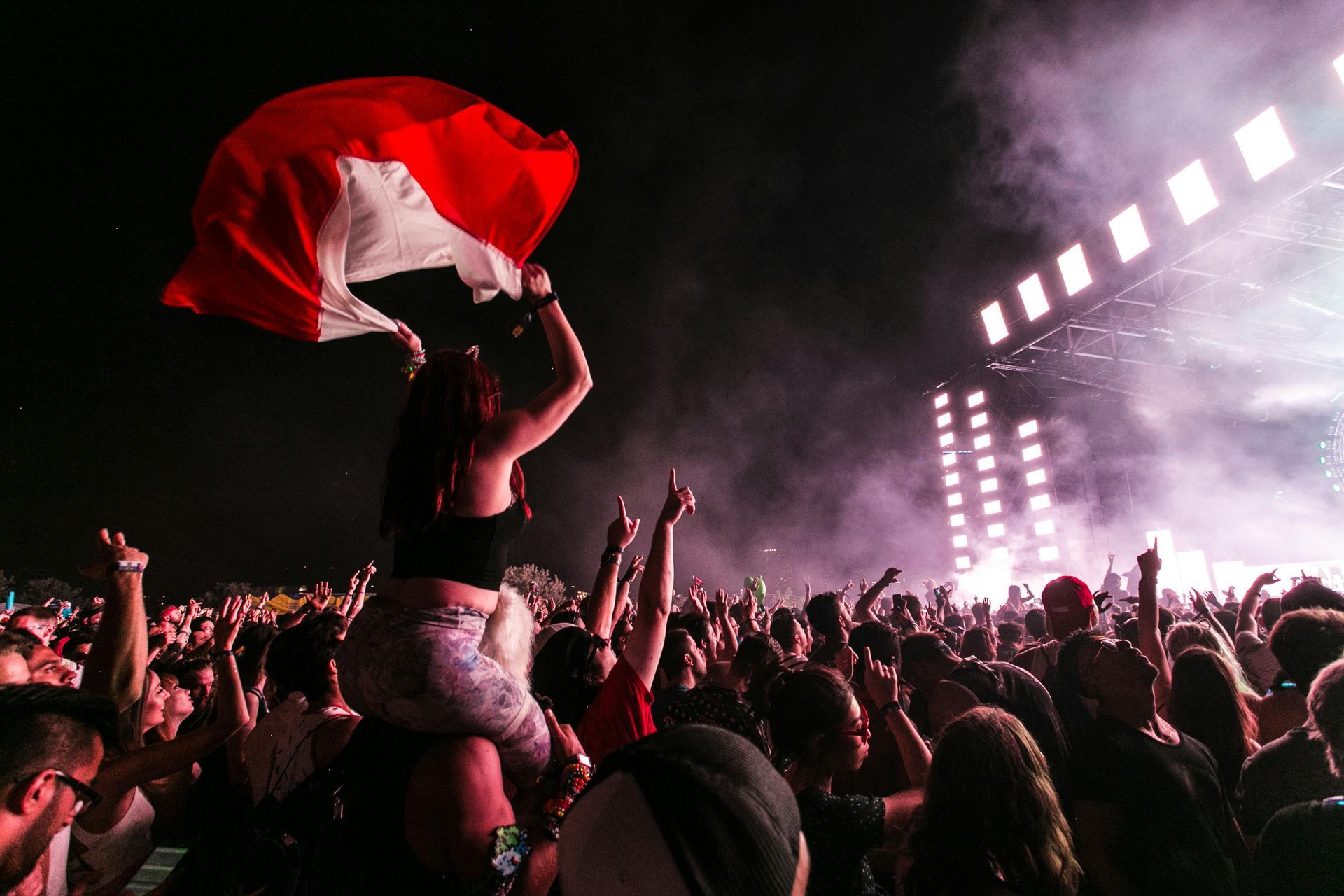 A woman on shoulders waves a red and white flag at a concert with a bright stage and cheering crowd.