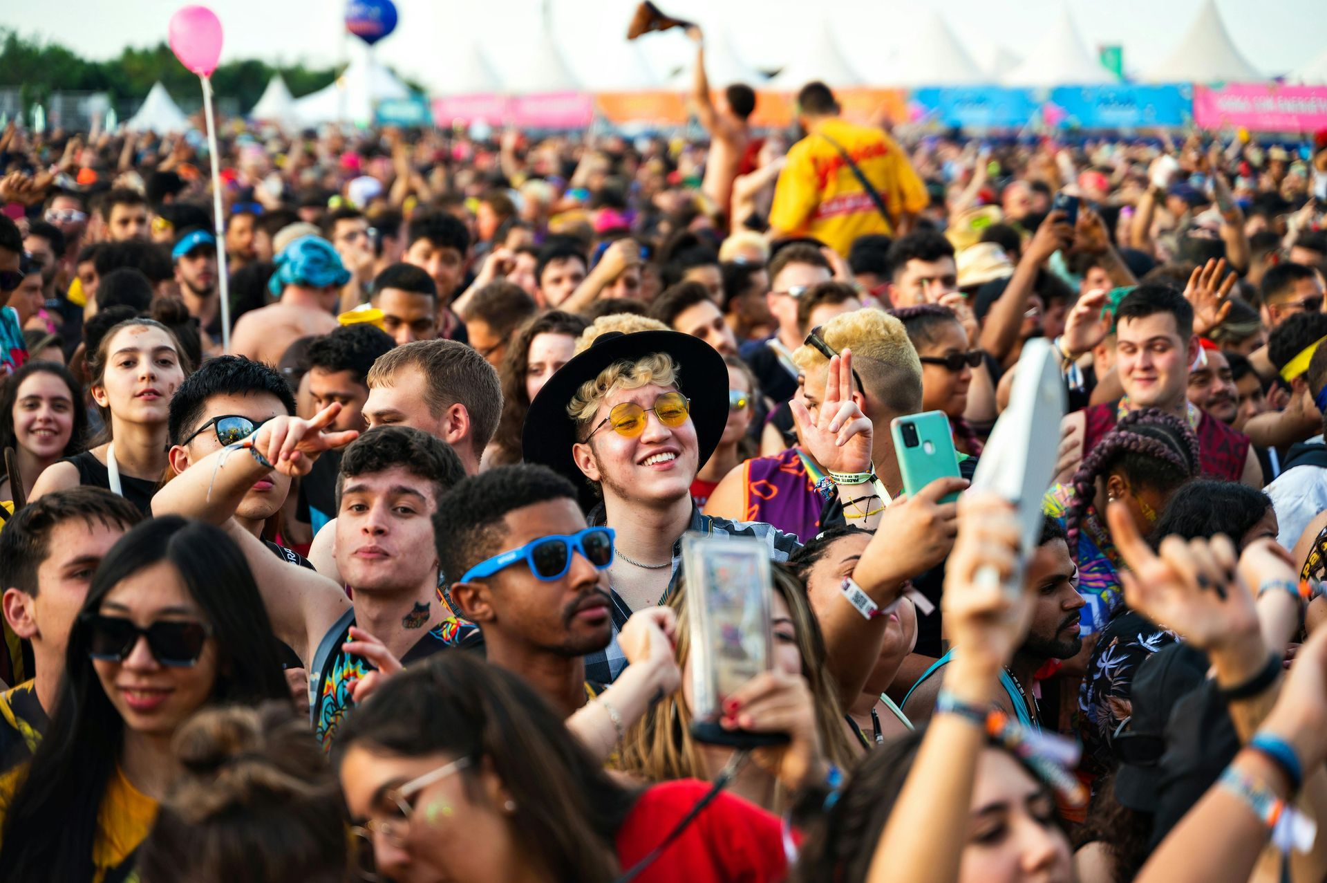 Large crowd at an outdoor music festival; many people, some holding phones and waving, with tents in the background.
