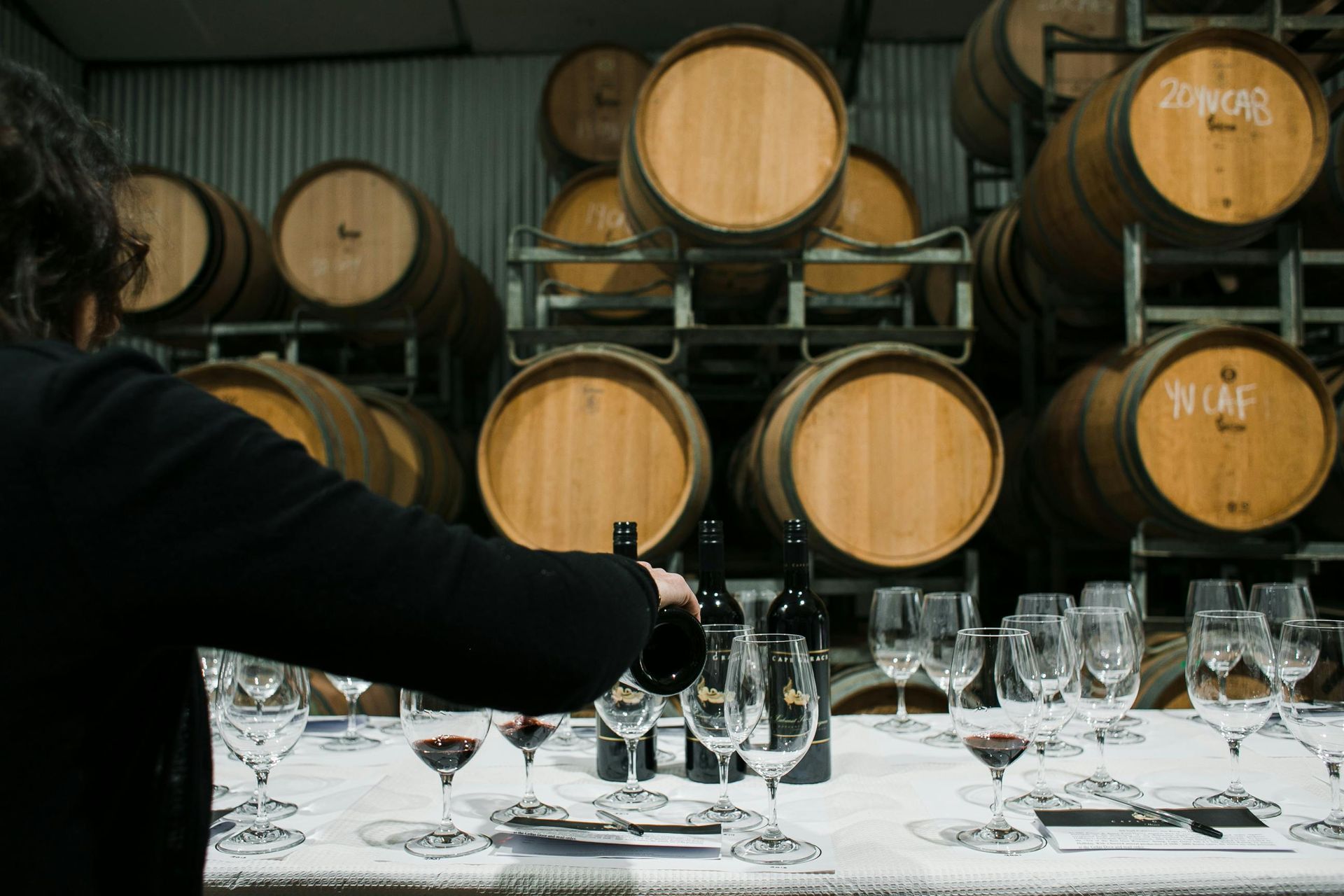 Person pouring wine at a tasting; barrels in background, empty glasses on table.