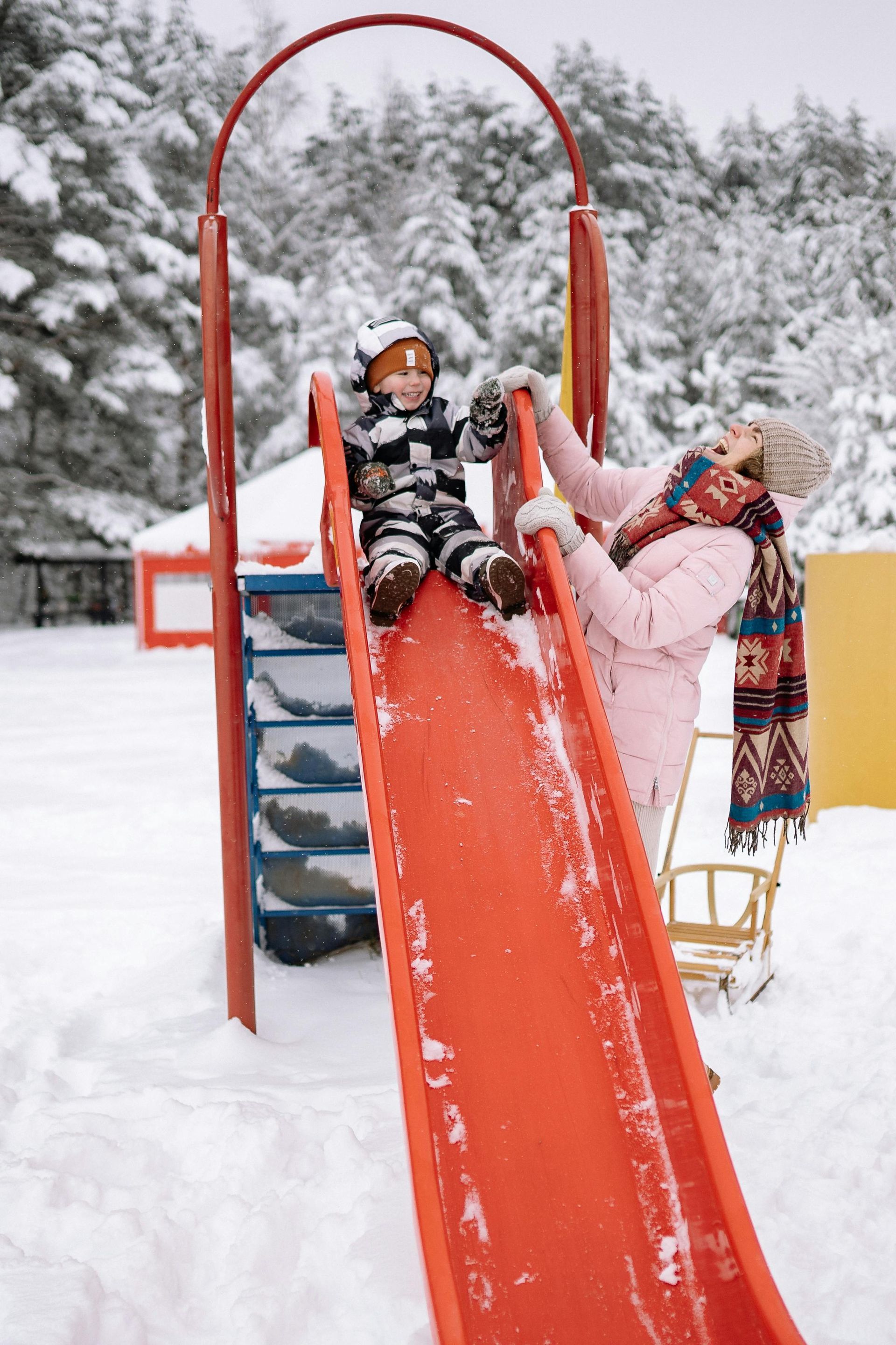 Woman assists child sliding down an orange slide covered in snow, in a winter playground.