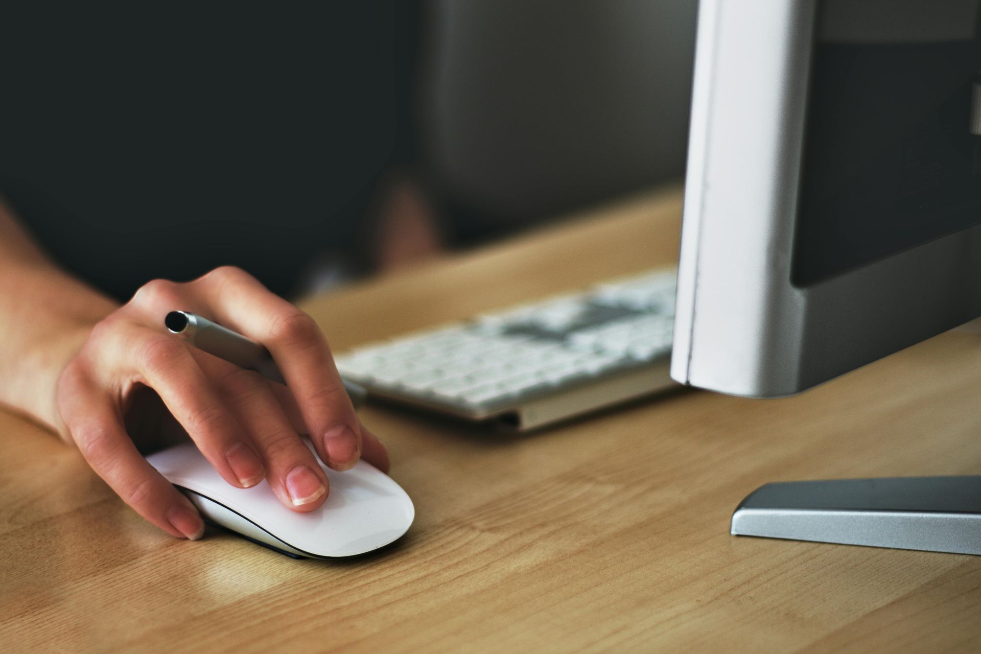 Hand holding computer mouse next to keyboard and monitor on a wooden desk.