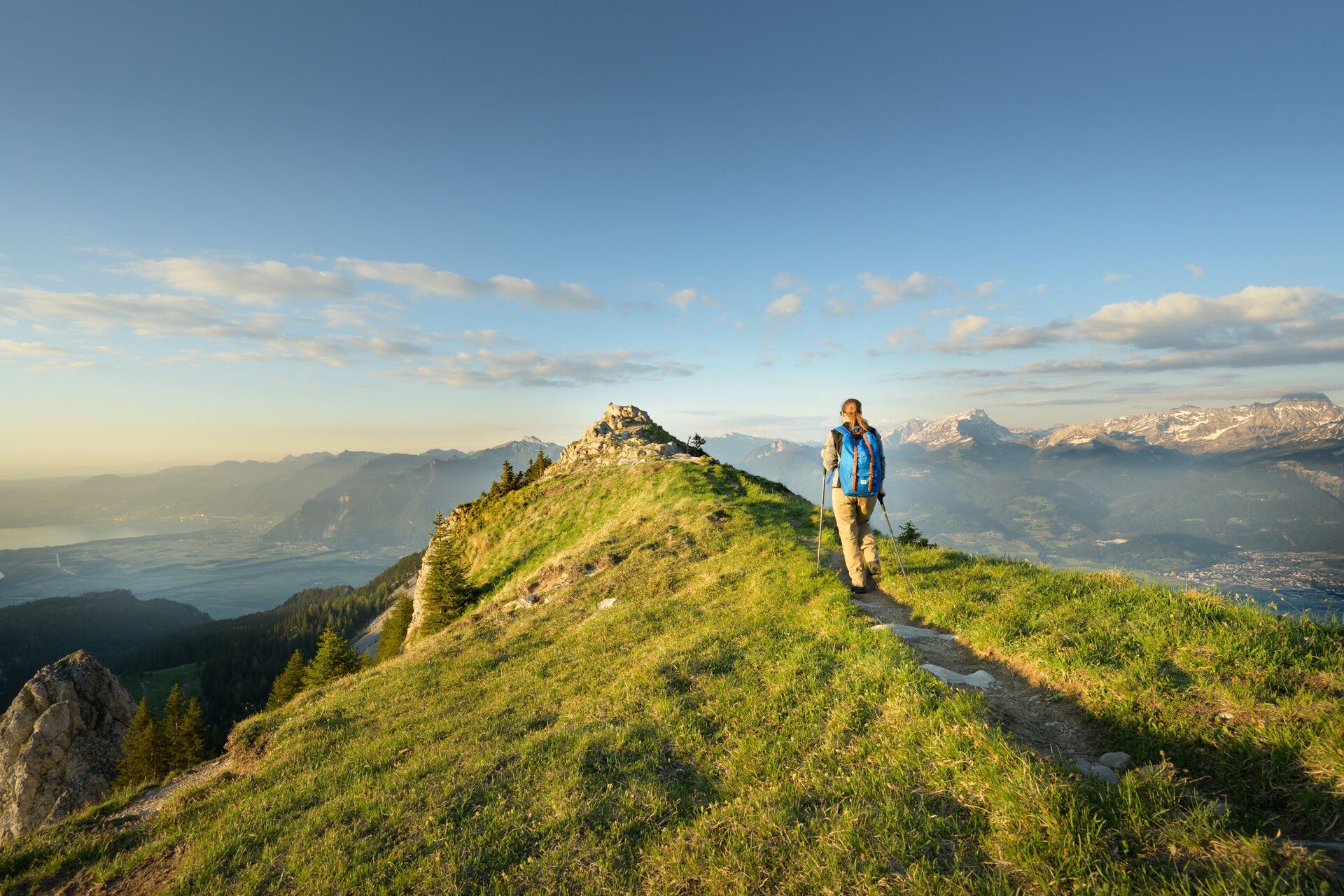 Hiker on a grassy ridge overlooking mountains at sunrise with a blue sky and distant clouds