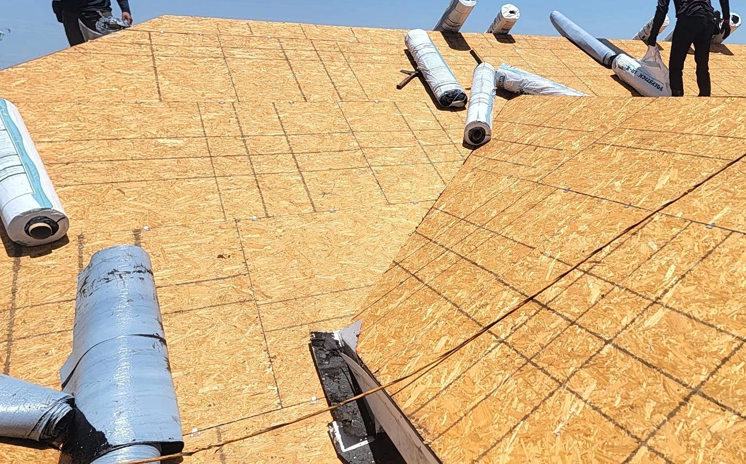 A man is standing on top of a roof with rolls of plywood on it.