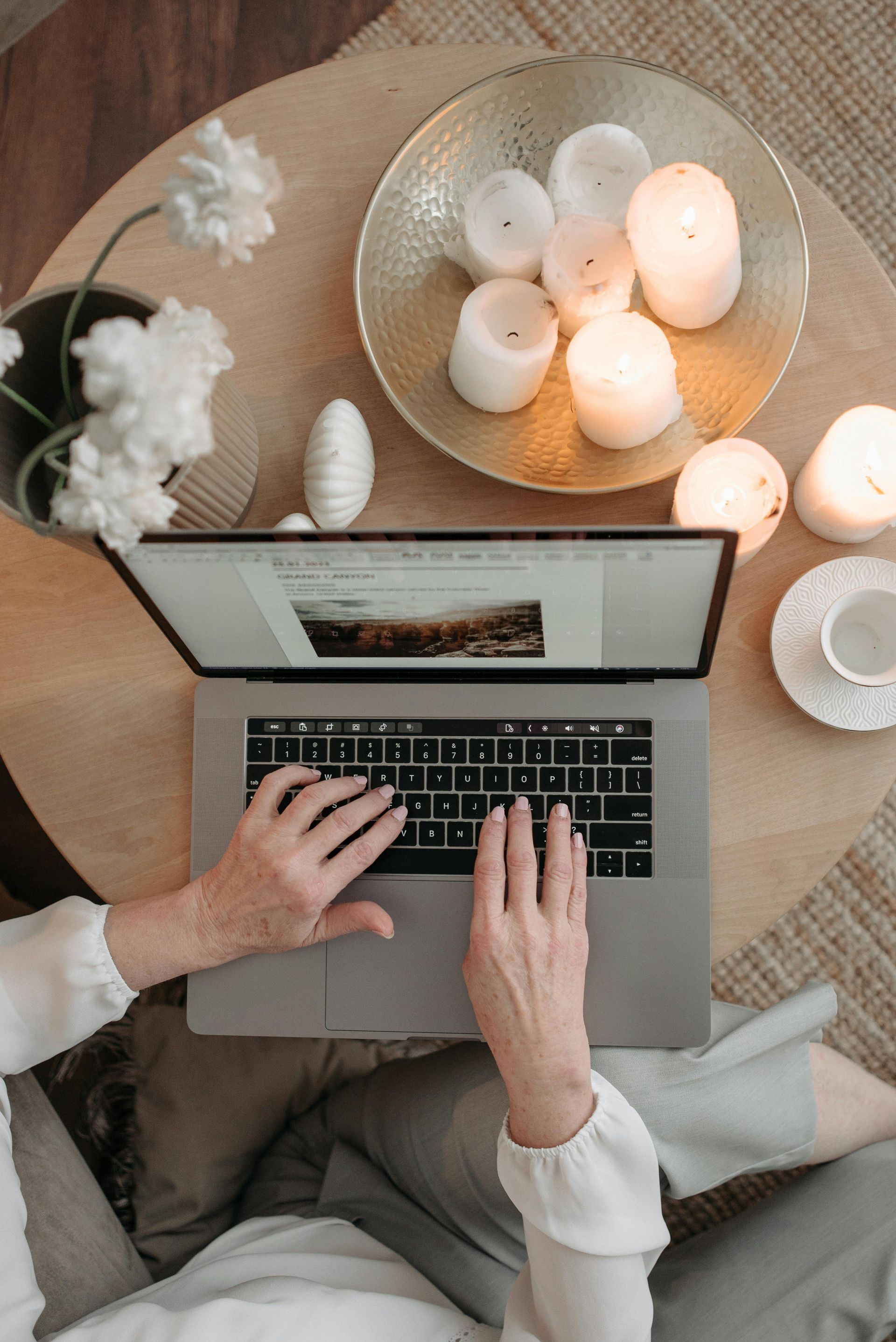 Hände tippen auf einem Laptop an einem mit Kerzen beleuchteten Tisch, neben dem Blumen und eine Tasse stehen.