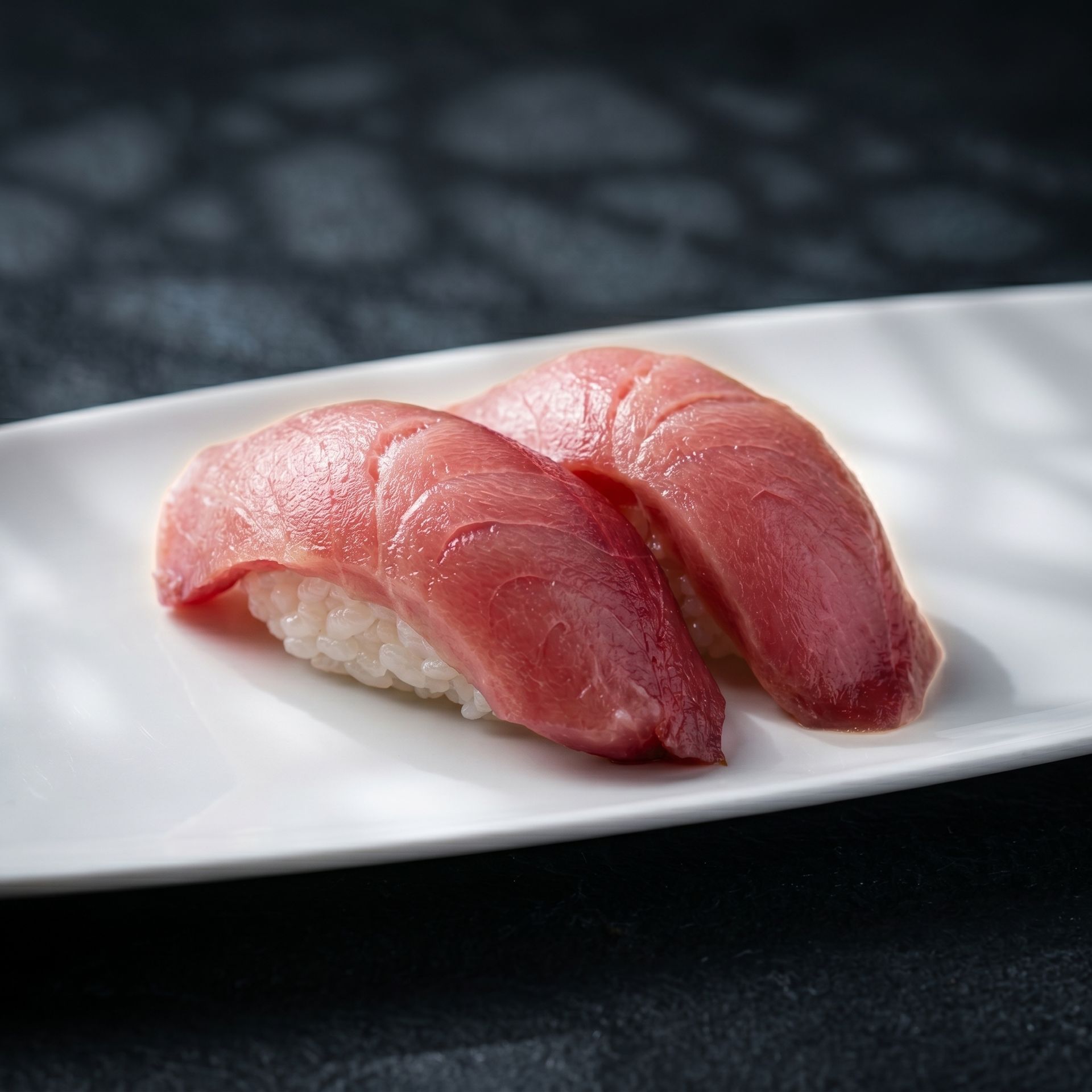 Two pieces of pink tuna nigiri on a white plate against a dark background