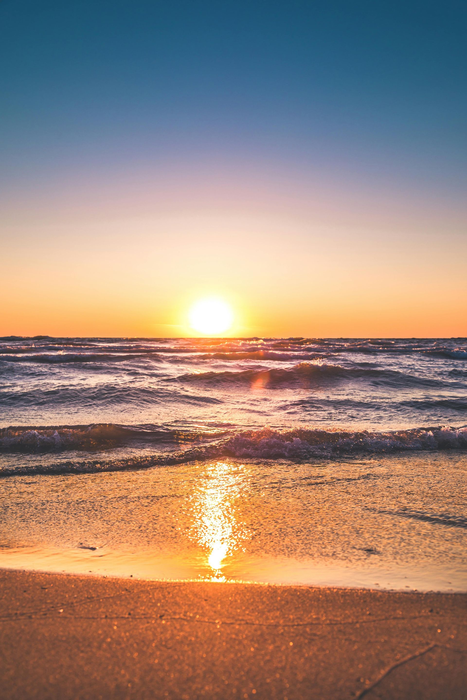 Sunset over the ocean with golden light reflecting on the wet sand and water; blue sky above.
