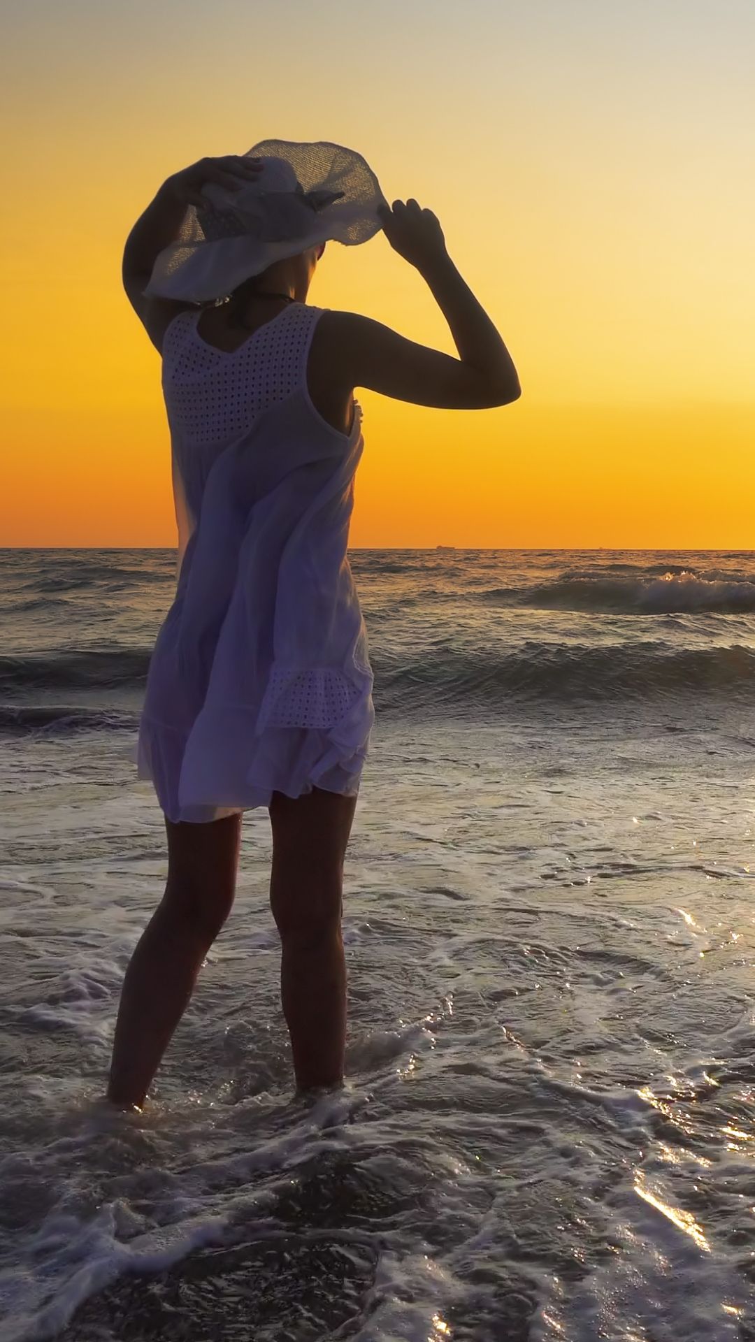 Woman in a white dress and sun hat standing in the ocean at sunset, silhouetted against an orange sky.