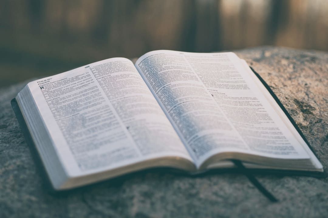 Open book with printed pages resting on a gray stone surface. A black ribbon bookmark is visible.