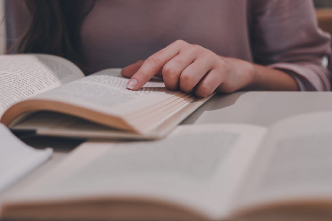 Person reading a book, pointing a finger at the text. Books are open on a table.