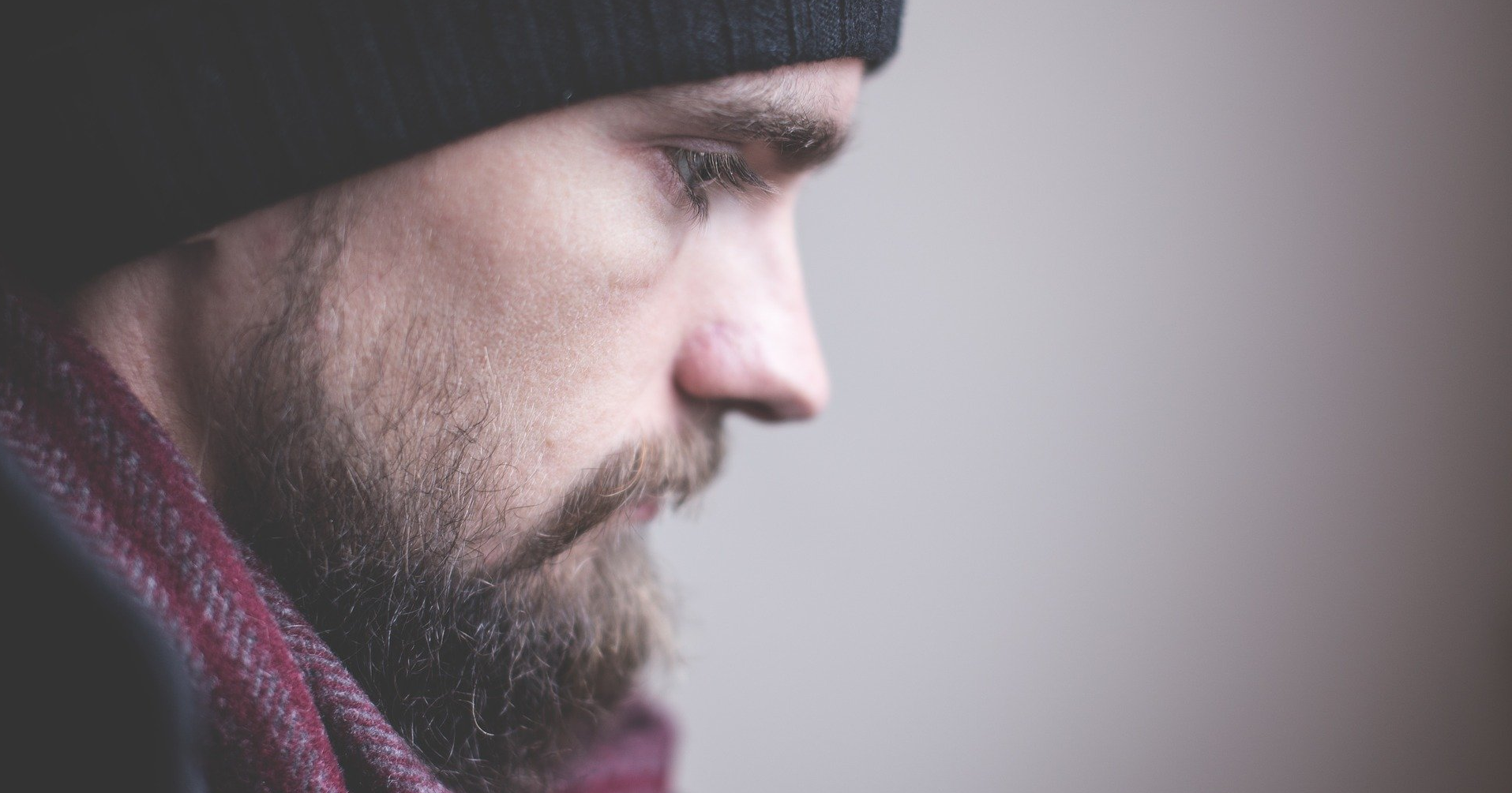 a man with a beard wearing a hat and scarf is looking down with emotional sadness.