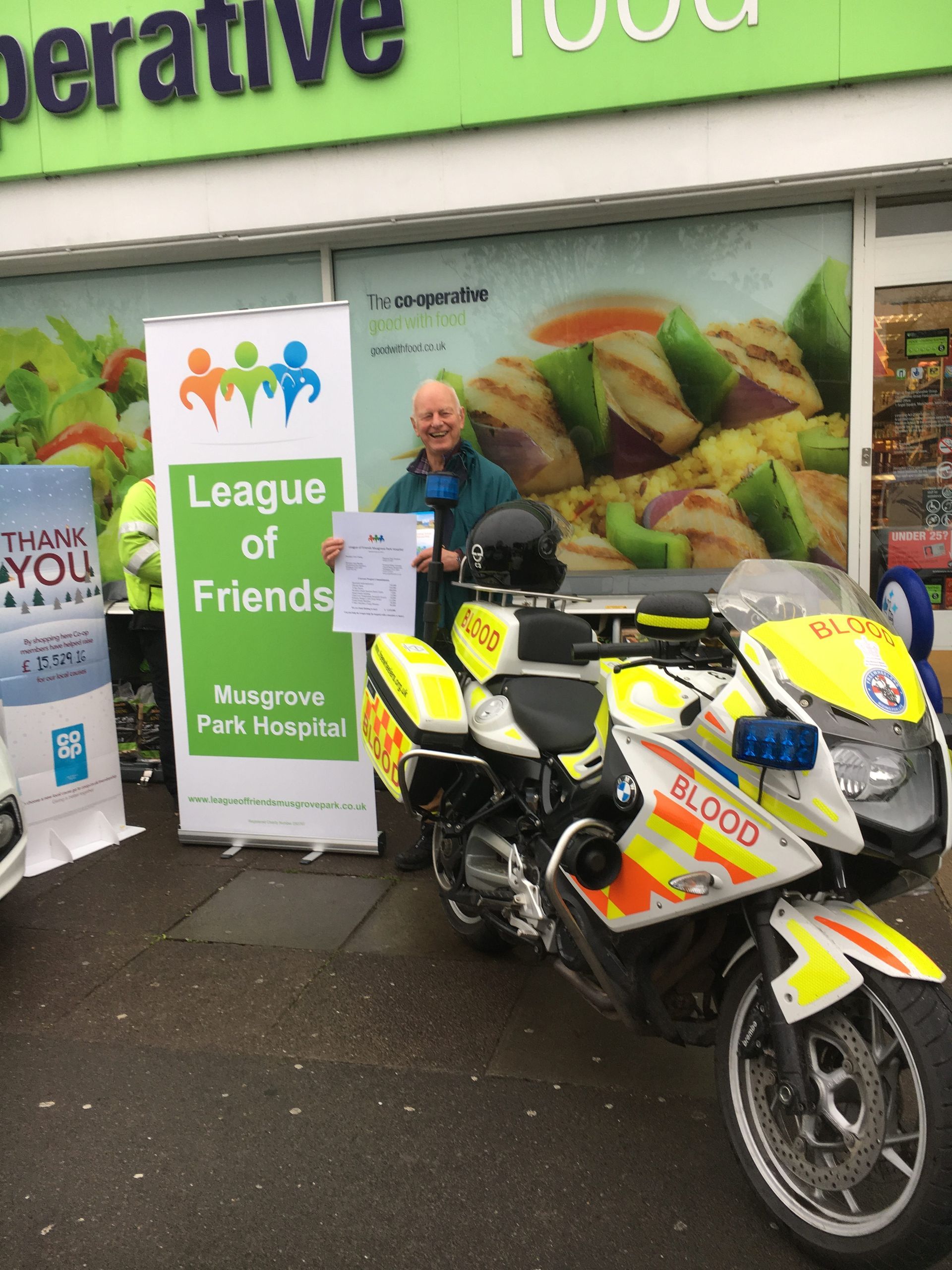A man standing next to a motorcycle in front of a sign that says league of friends, fundraising.