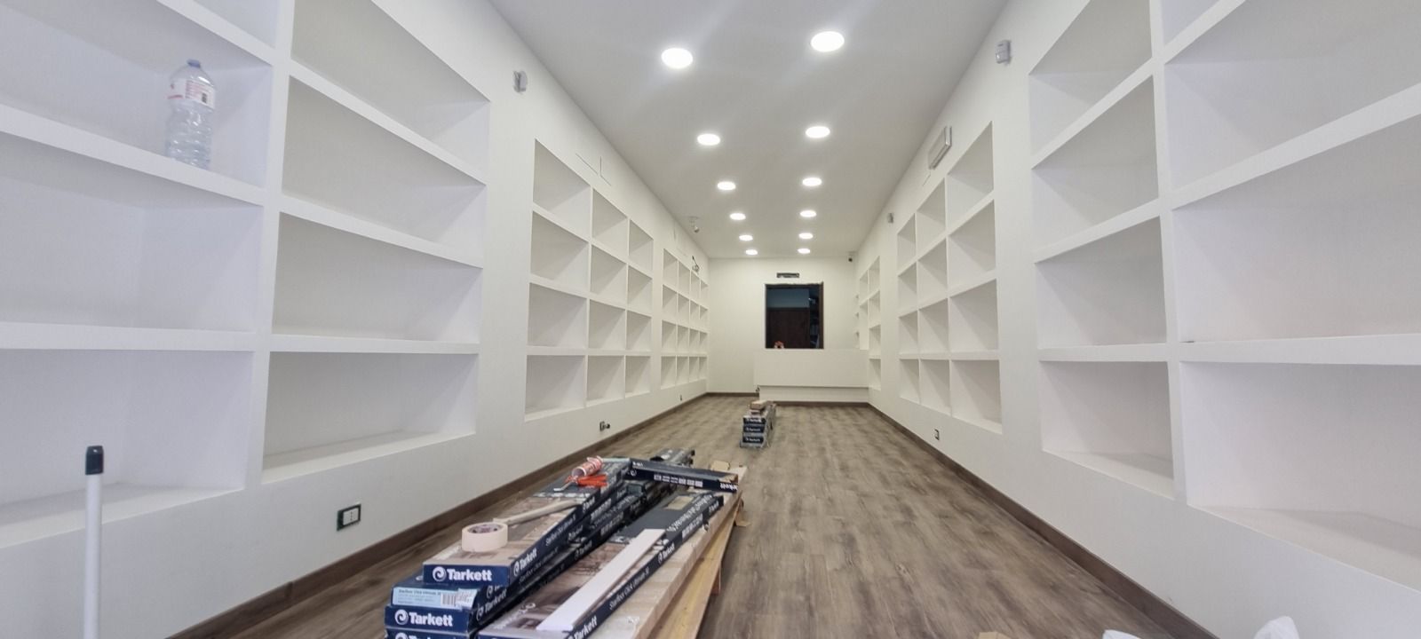 Una lunga stanza con scaffali bianchi a muro. Il pavimento è color legno e in fondo si vede una piccola finestra.