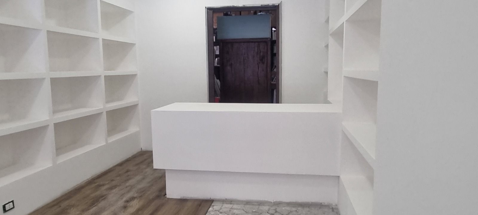Uno spazio commerciale bianco con scaffalature integrate, un bancone bianco e una porta d'ingresso marrone scuro.