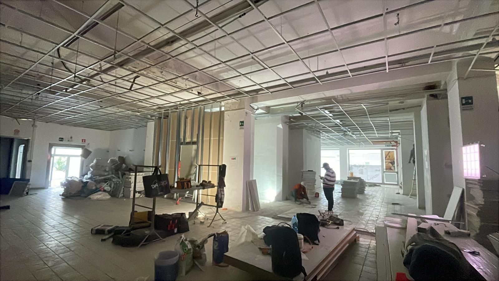 Cantiere interno: soffitto in griglia metallica, operai, travi a vista, materiali, spazio luminoso e aperto.
