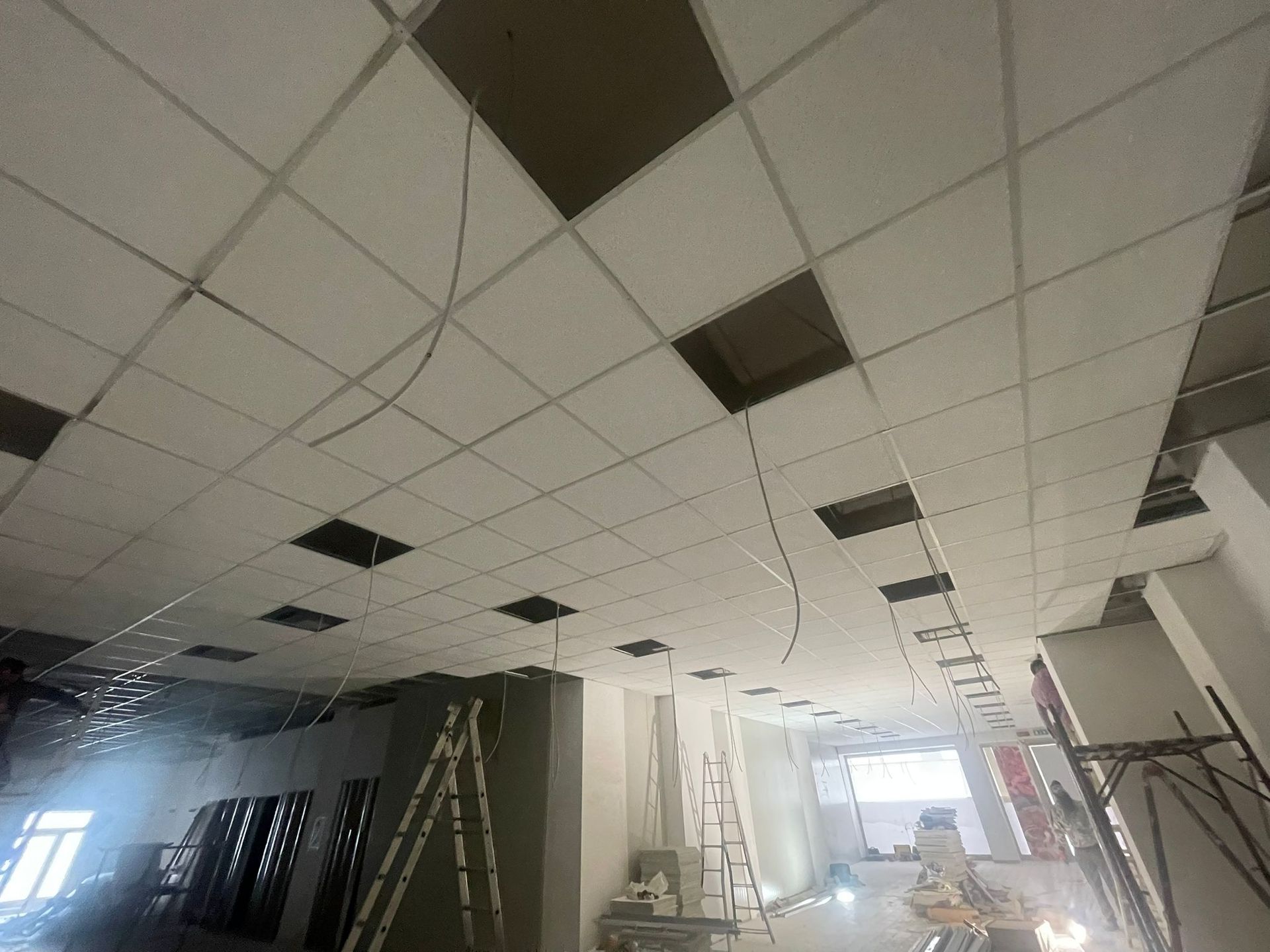 Interno di un edificio in costruzione. Controsoffitto con piastrelle mancanti; cavi elettrici esposti. Scale e materiali visibili.