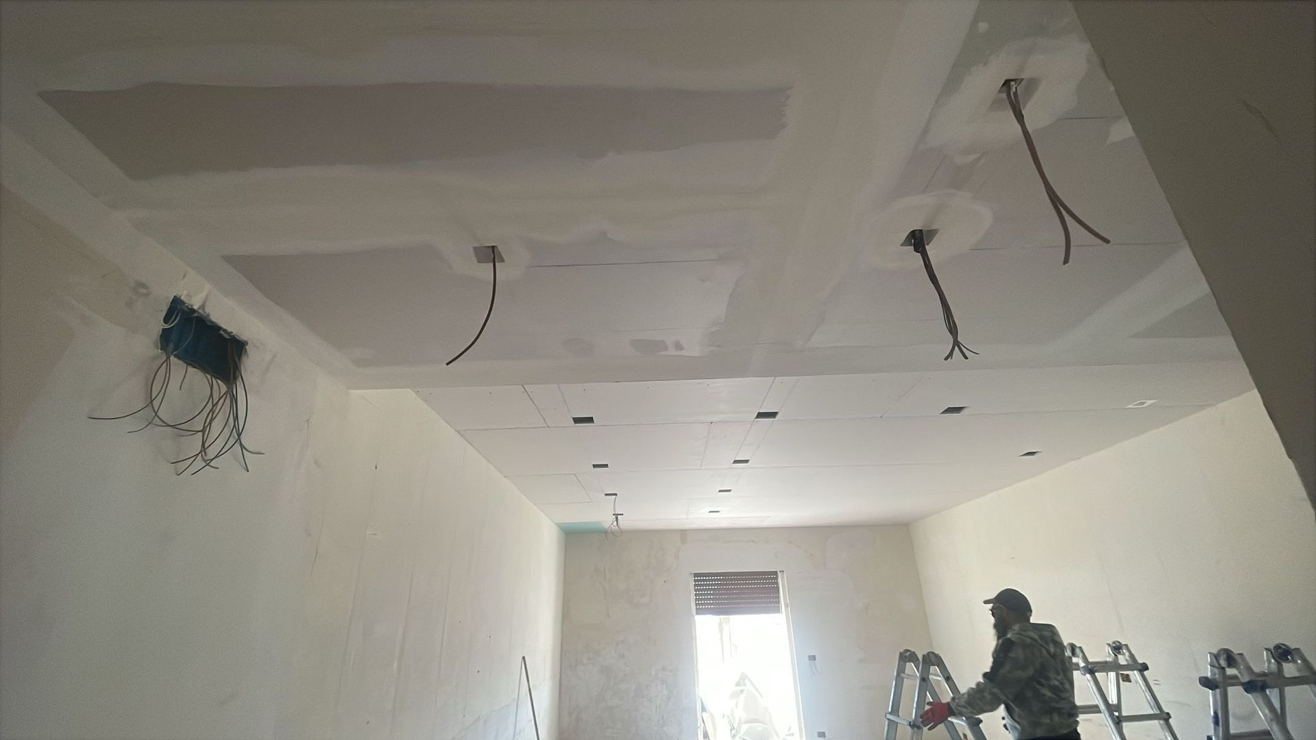 Stanza in costruzione; soffitto e pareti in cartongesso, fili elettrici scoperti e una persona su una scala.