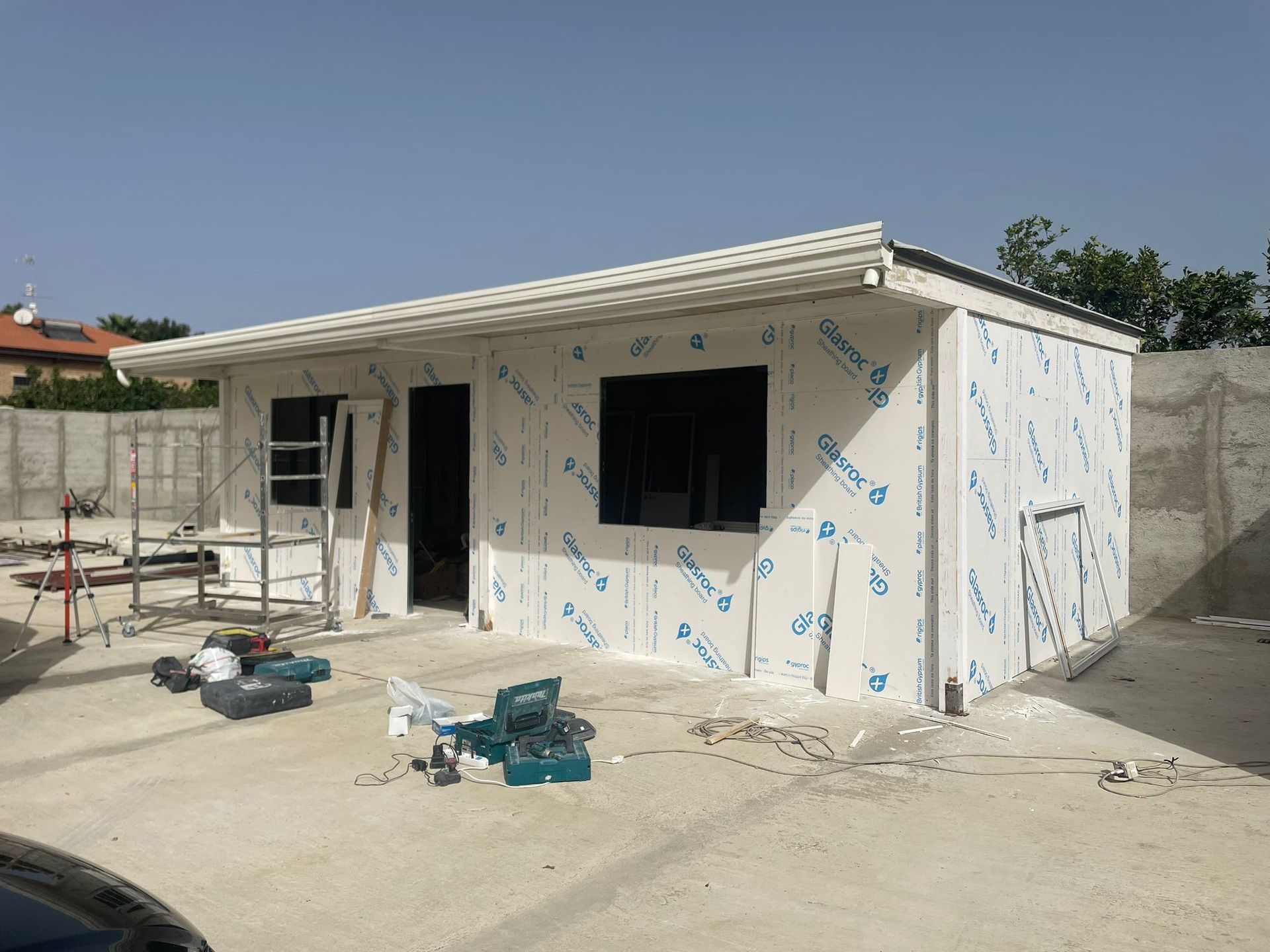 Un piccolo edificio in costruzione; pareti rivestite di pannelli bianchi, infissi neri, attrezzi sparsi su una superficie di cemento, cielo azzurro.
